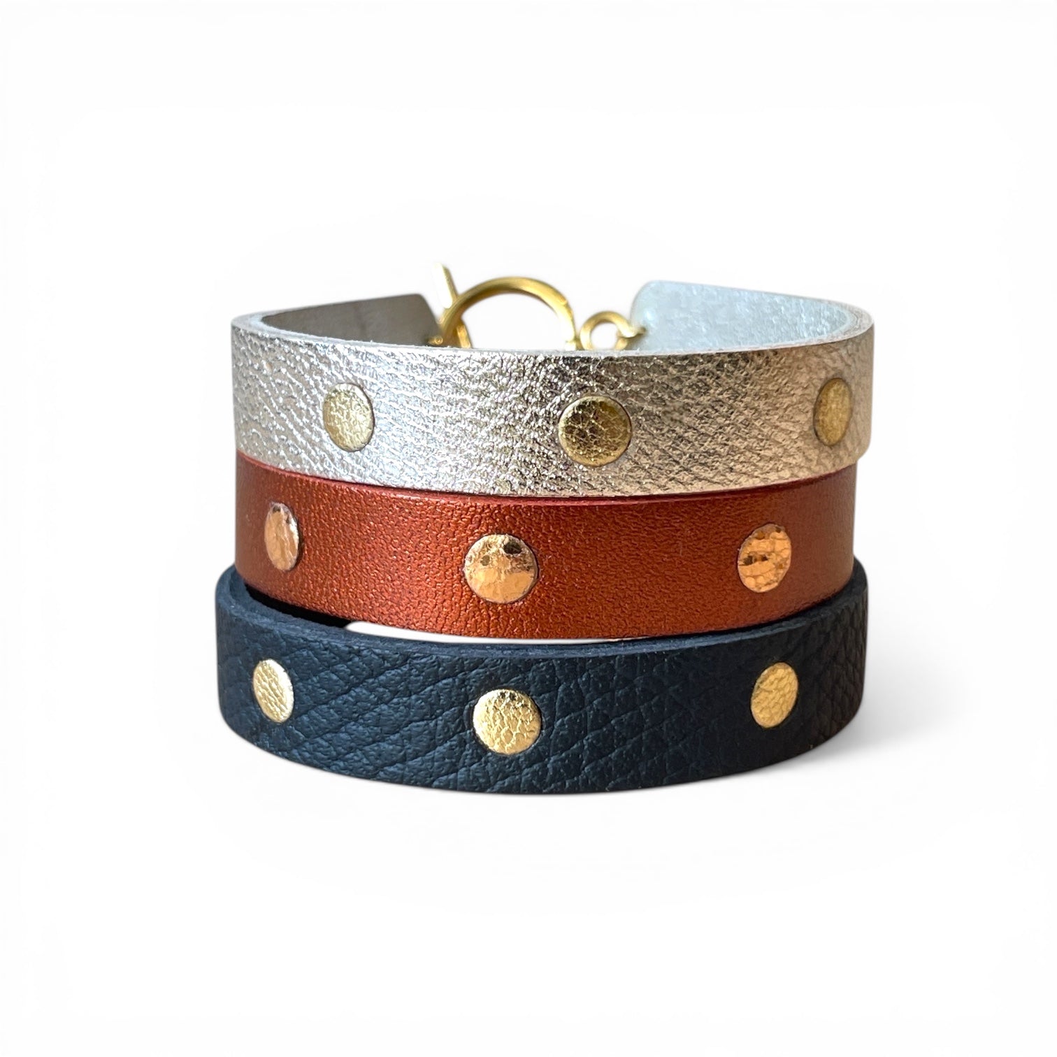 3 Bracelets dorés en cuirs bicolore argent et or, terracotta métallisé et or rosé froissé, et noir grainé et or, empilés en tour les uns sur les autres, sur fond blanc