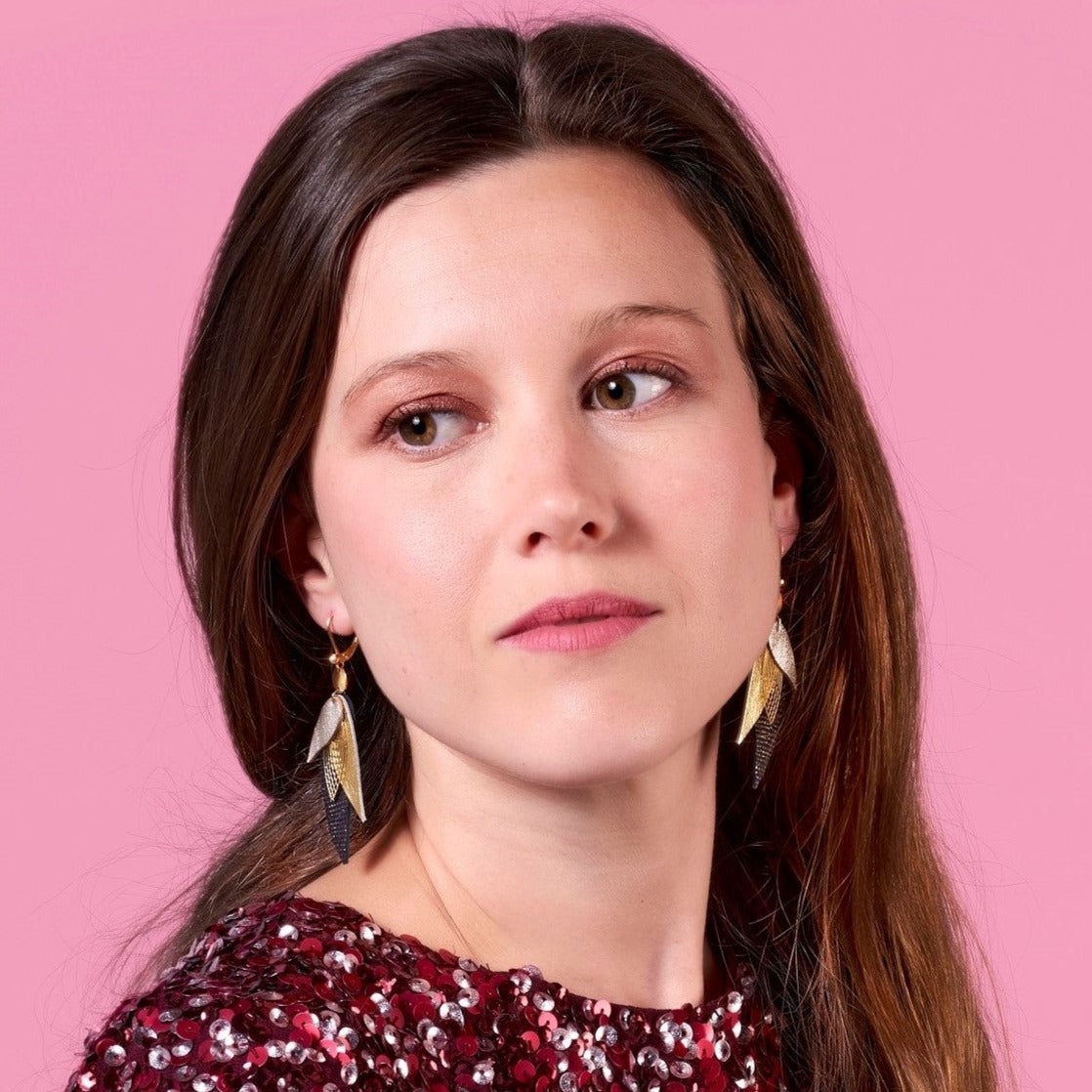 Jeune femme aux longs cheveux bruns portant les boucle d'oreille dorée Nune modèle Ara en plumes de cuir argent, doré brillant et noir grainé irisé, sur fond rose