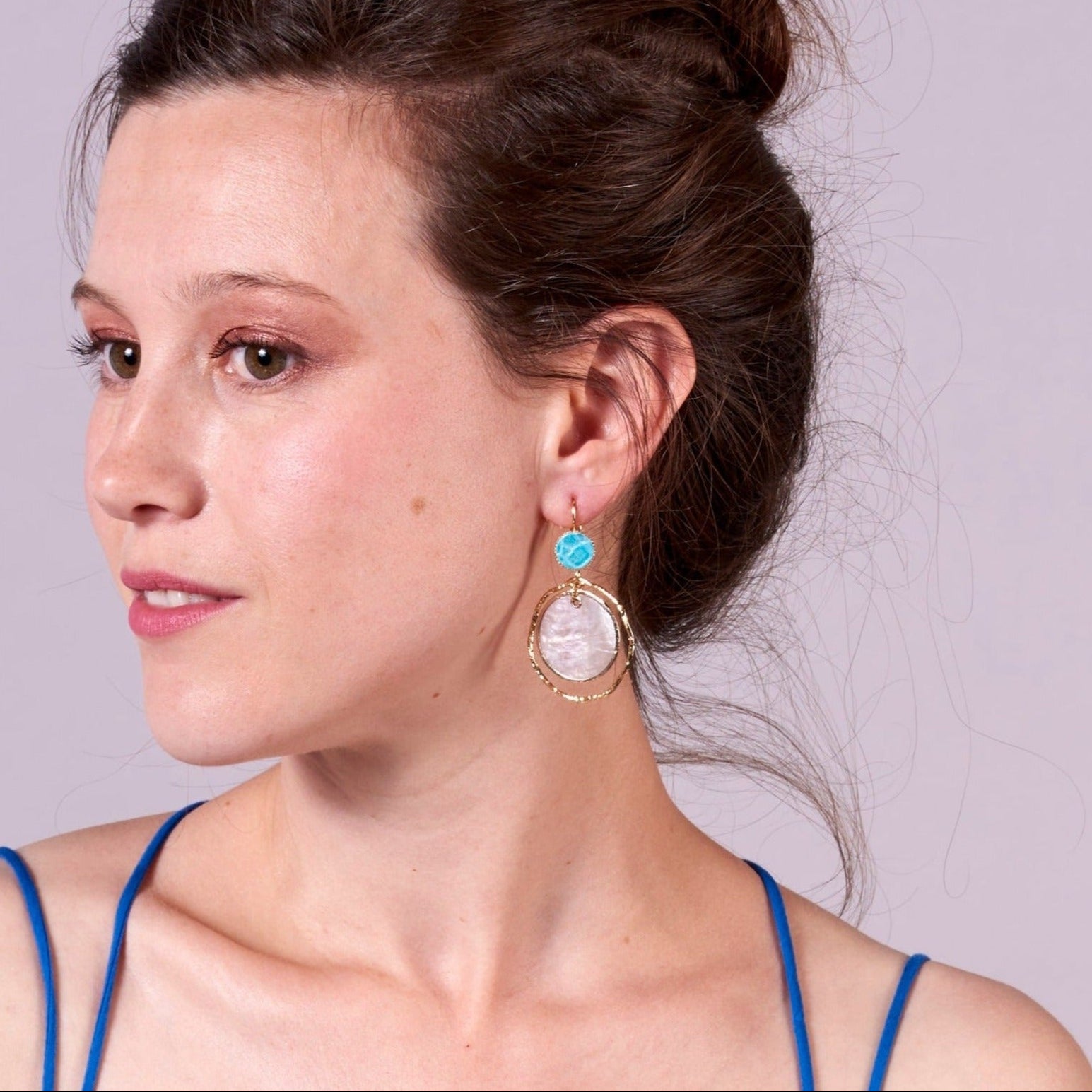 Jeune femme à chignon vue de profil portant les boucles d'oreilles dorées Nune modèle Bali Créoles Capiz en cuir turquoise sur fond