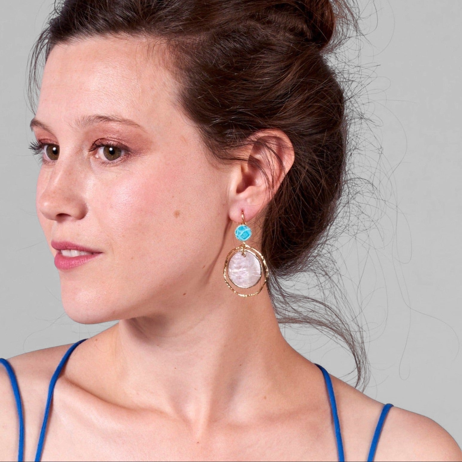 Jeune femme à chignon vue de profil portant les boucles d'oreilles dorées Nune modèle Bali Créoles Capiz en cuir turquoise sur fond gris