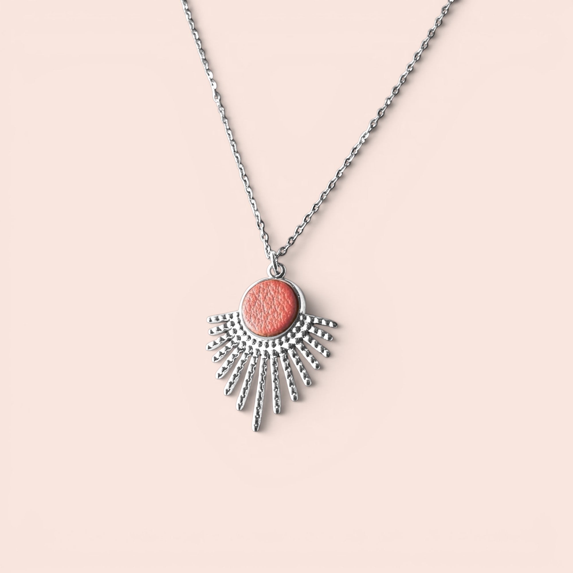 Collier argenté fin avec pendentif en forme de petit soleil, avec cabochon de cuir coloré corail nacré, sur fond rose pâle