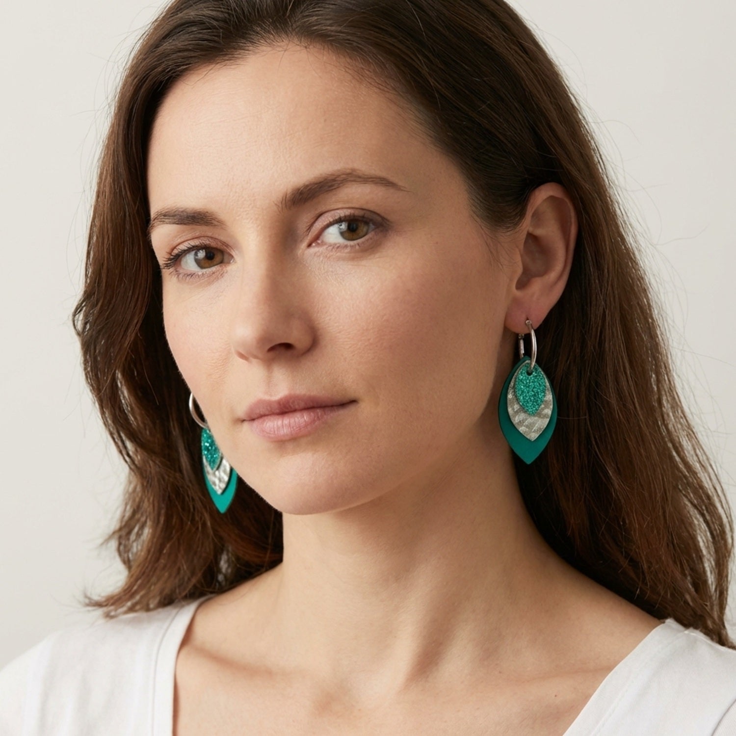 Créoles argentées avec pendentifs modulables en forme de feuilles en cuirs vert émeraude métallisé, argent grainé et vert émeraude grainé, portées à l'oreille par une femme brune