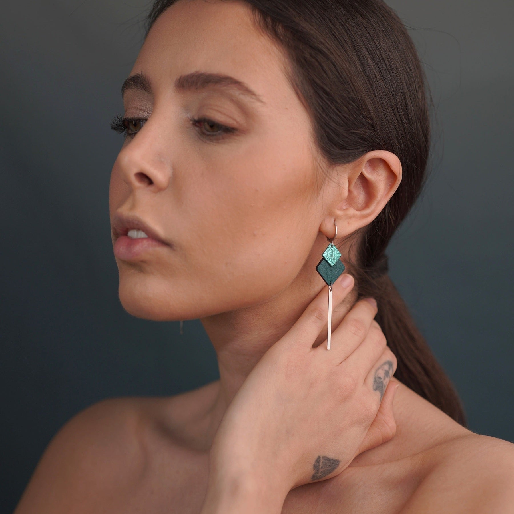 boucles d'oreilles argentées Nune modèle Ada Argenté avec duo de losanges en cuirs vert canard et vert émeraude brillant et longue tige fine argentée, portées à l'oreille par une femme brune