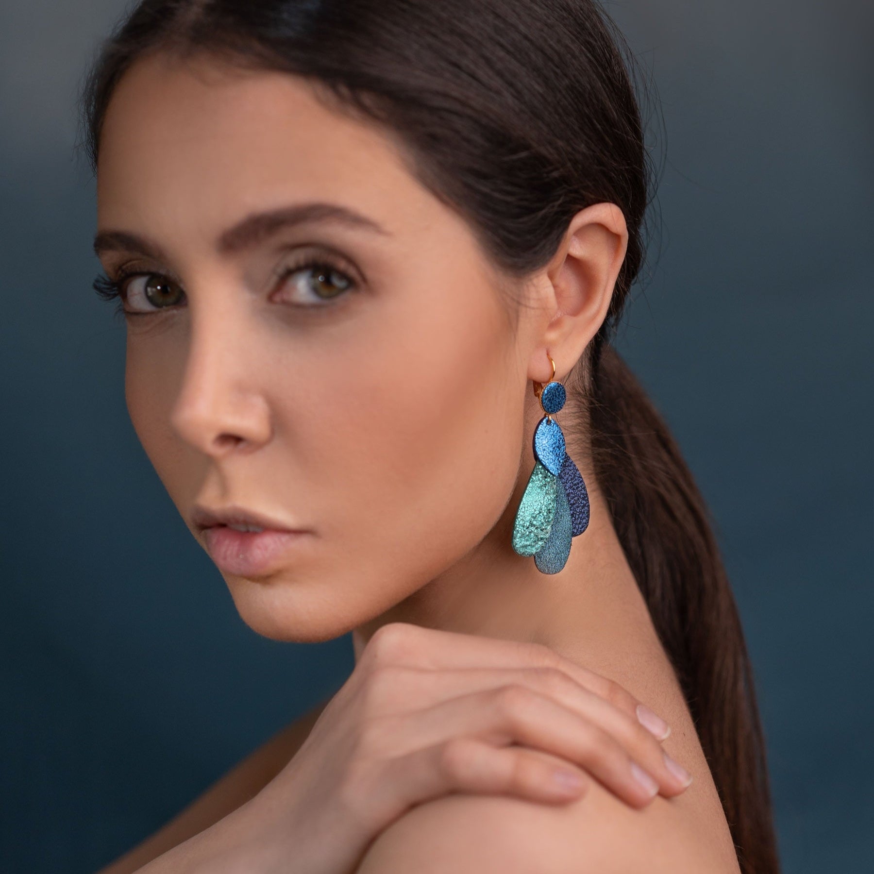Paire de grandes boucles d'oreilles dorées en forme de plumes de paon, en cuirs recyclés bleu roi métallisé, vert émeraude brillant intense, bleu paon pailleté et bleu marine pailleté, porté à l'oreille. Modèle Paon de Nune.