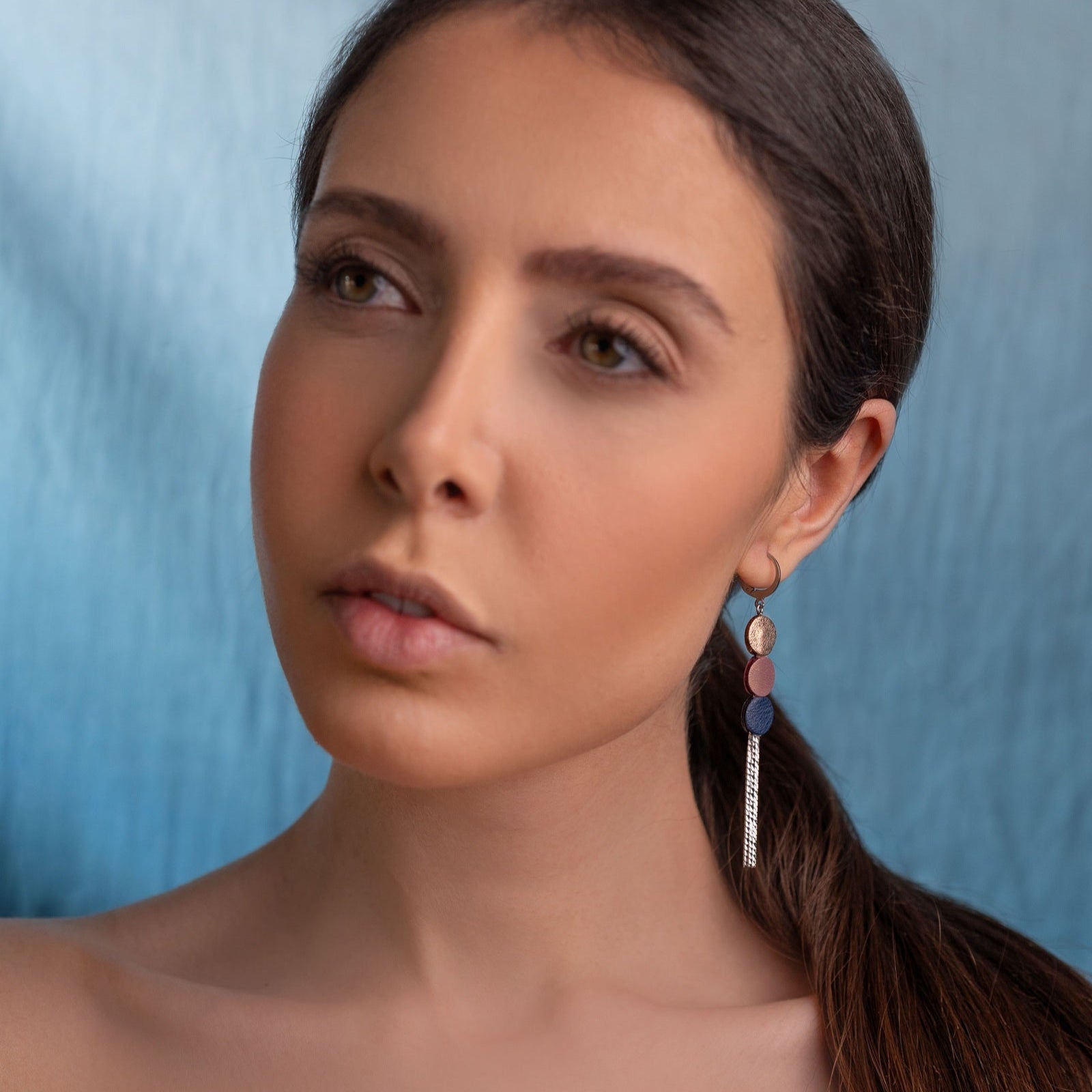 Longue et fine boucle d'oreille à franges argentées avec 3 petits disques de cuirs cannelle dorée satinée, vieux rose nacré et bleu marine, portée à l'oreille par une femme brune