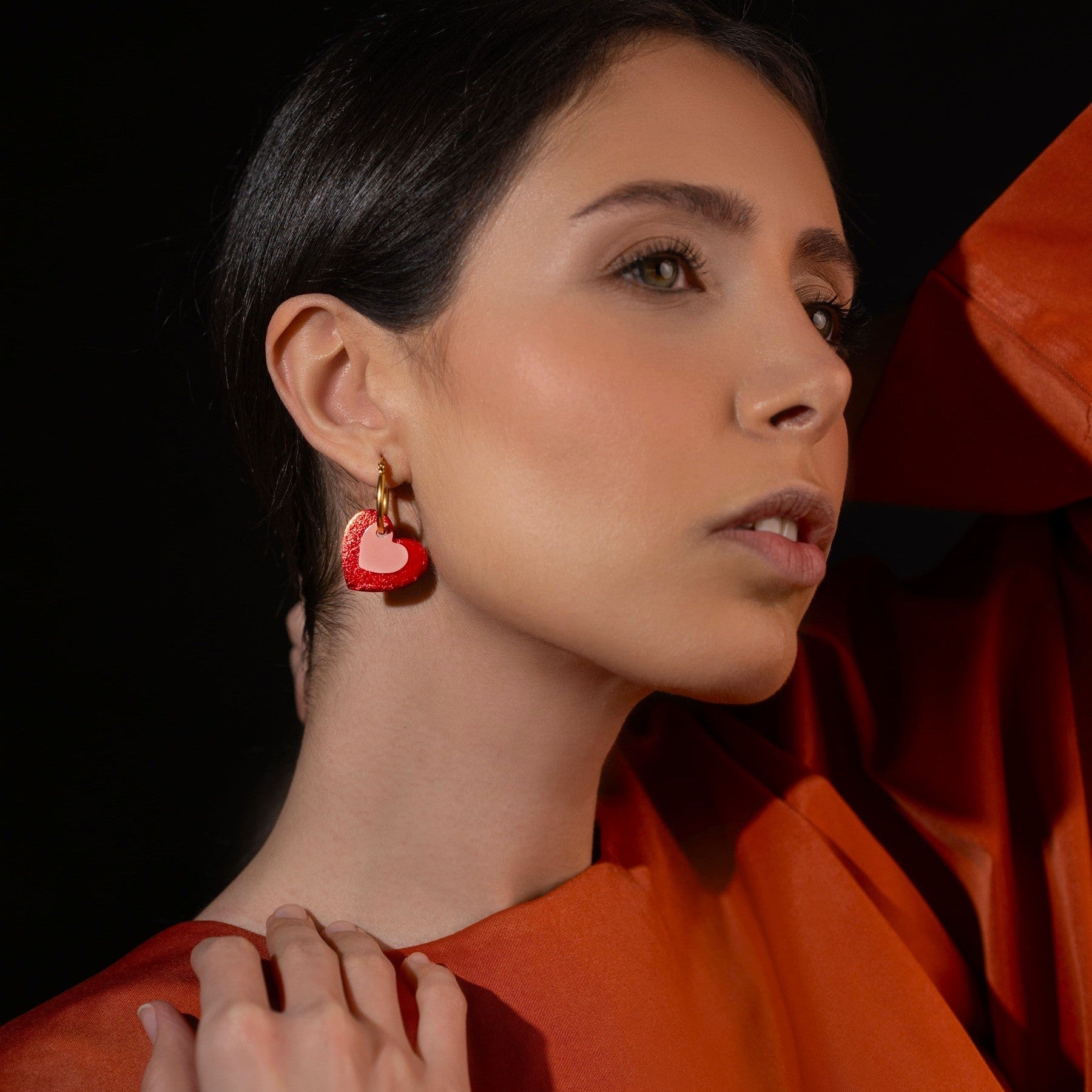 Boucles créoles dorées avec pendentifs en forme de cœurs rouge métallisé et rose vernis, portées à l'oreille par une femme brune