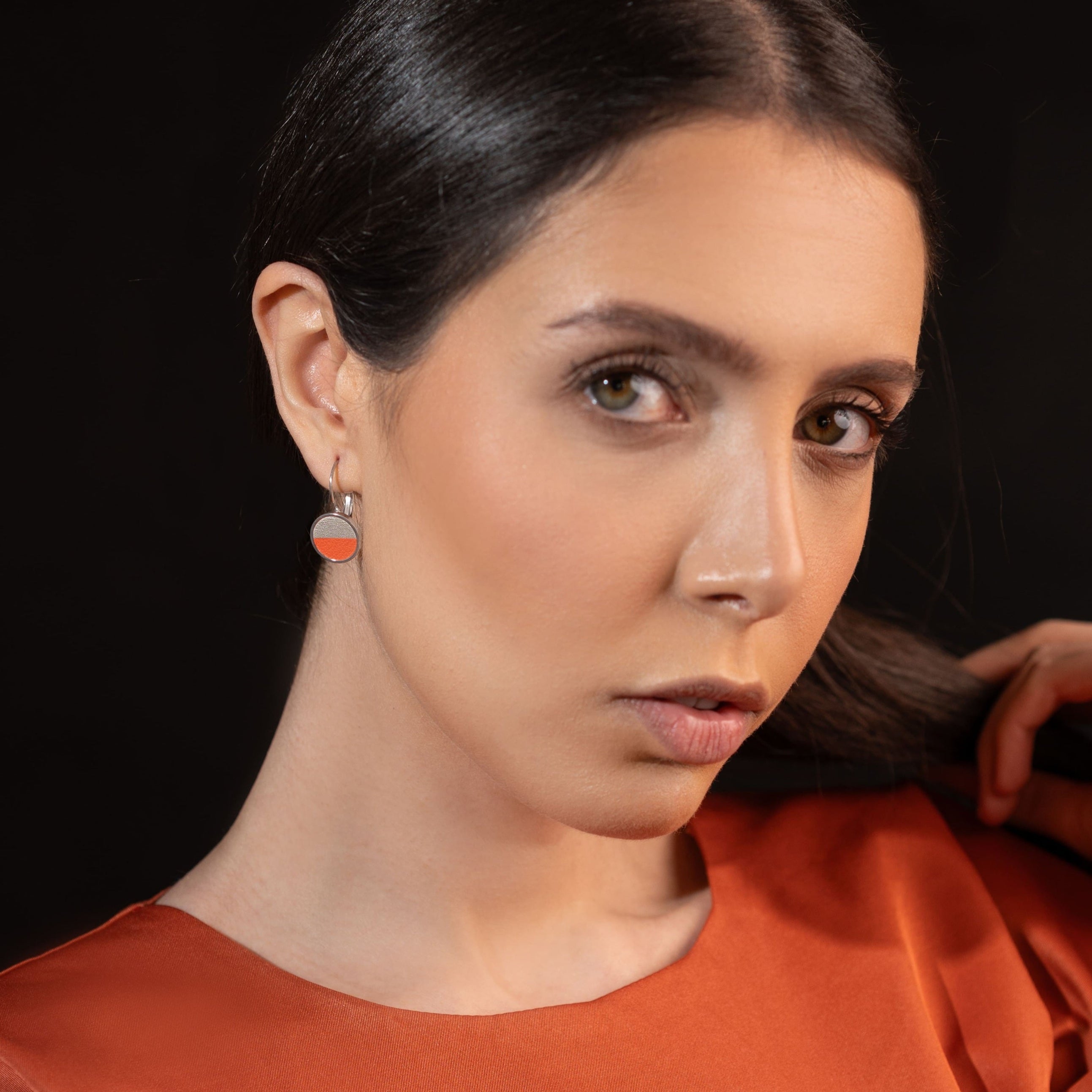 Petites boucles d'oreilles argentées pendantes rondes avec un duo de cuirs champagne nacré et carotte en cabochon, portées à l'oreille par une femme brune