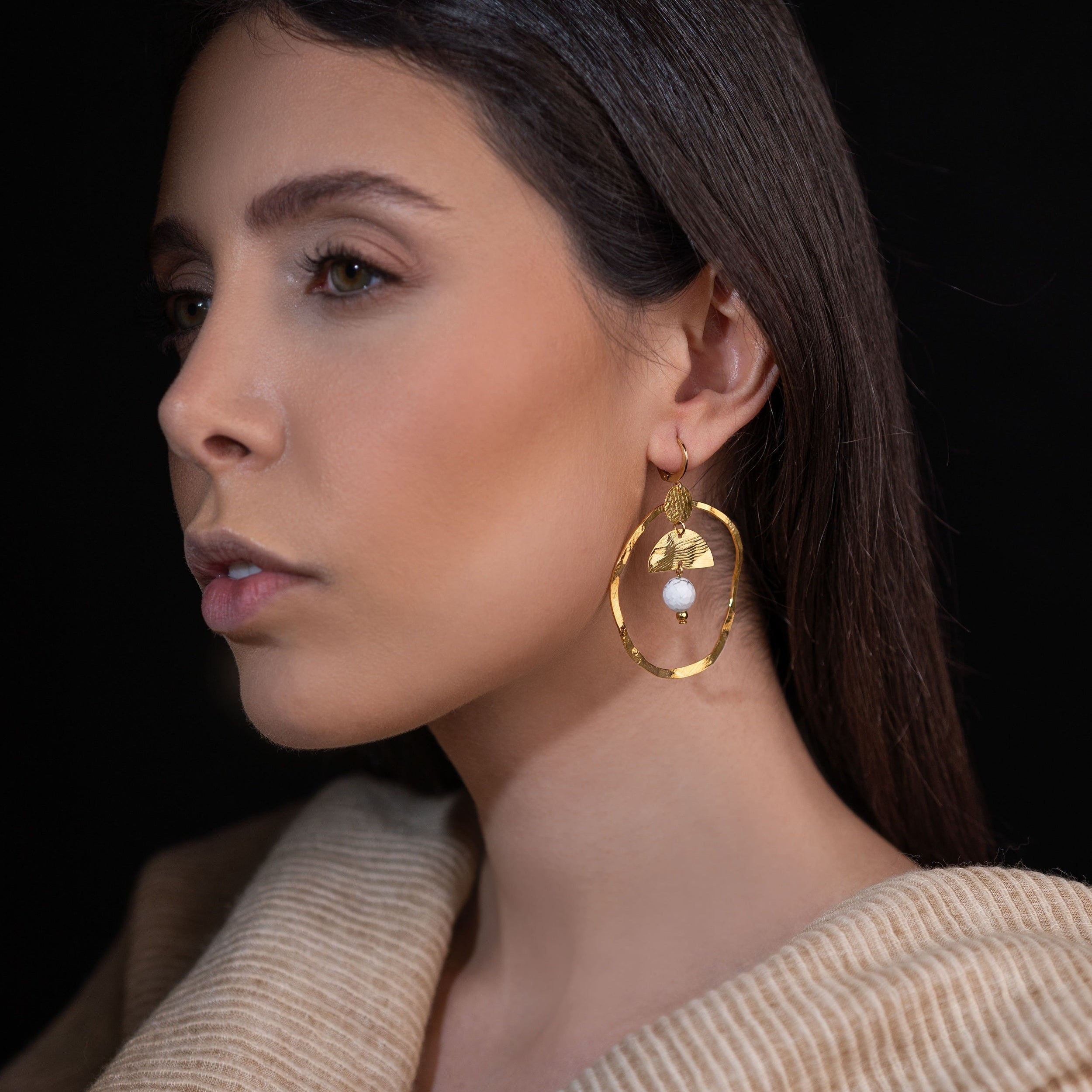 Grandes boucles d'oreilles créoles en métal martelé doré à l'or fin et pierres naturelles howlite blanche accrochée à une demi-lune, portées à l'oreille par une femme brune