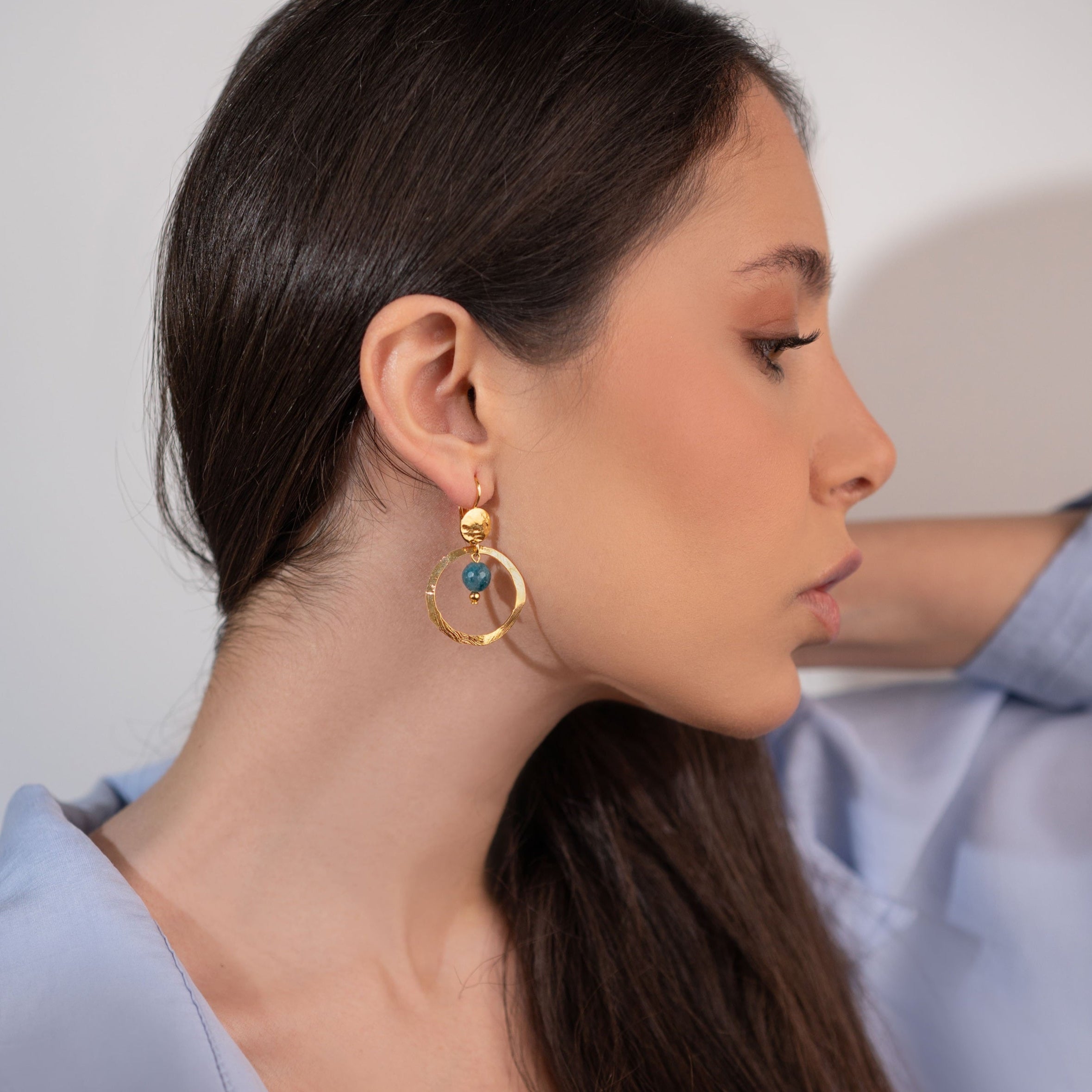 Petites boucles d'oreilles créoles dorées pendantes rondes avec perles d'apatite bleue, portées à l'oreille par une femme brune