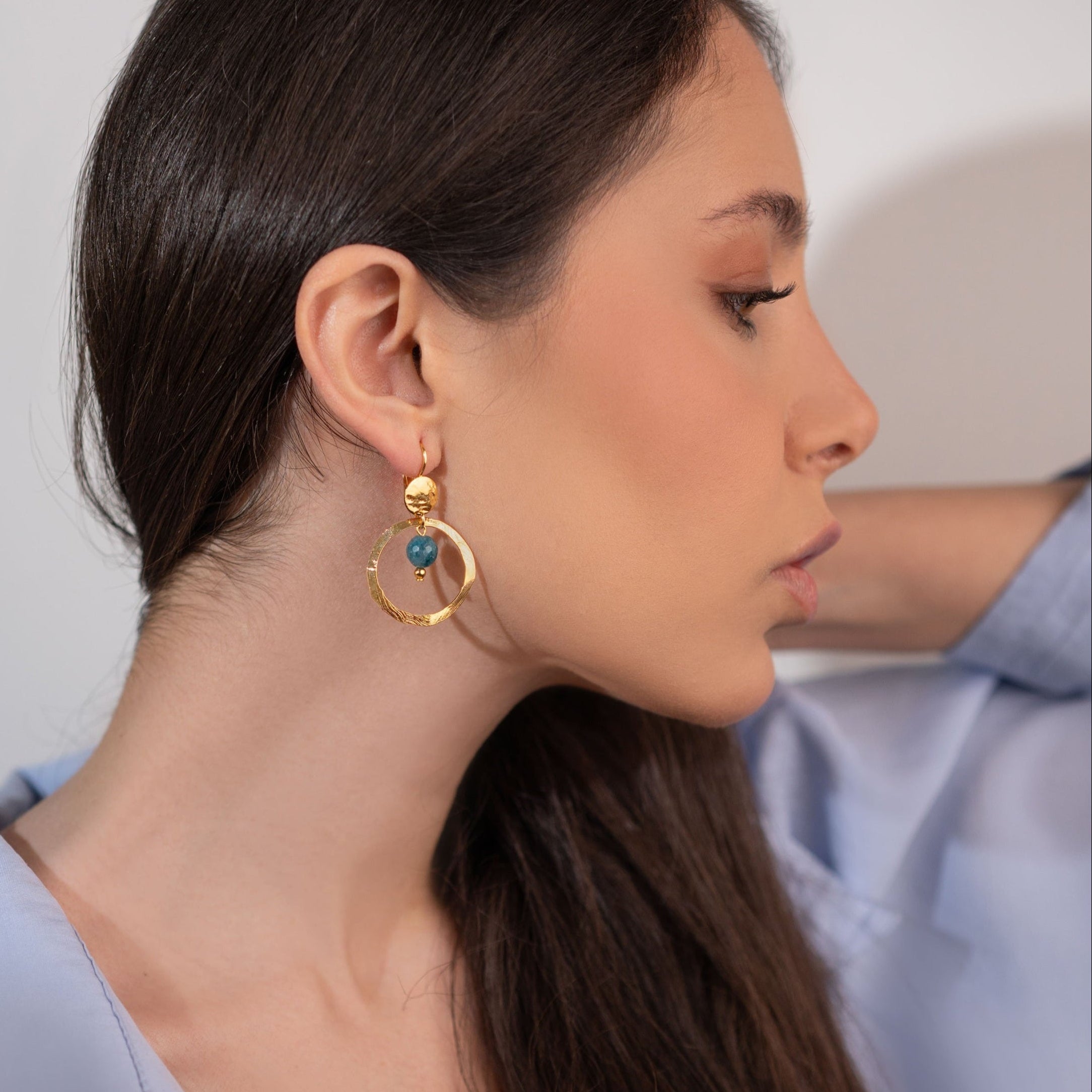Petites boucles d'oreilles créoles dorées pendantes rondes avec perles d'apatite bleue, portées à l'oreille par une femme brune