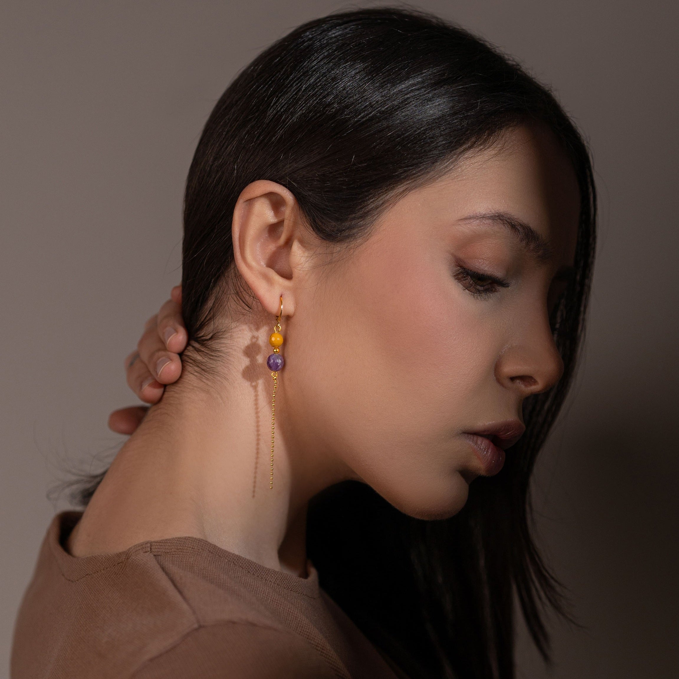 Fines boucles d'oreilles pendantes dorées en perles de pierres naturelles améthyste et mokaïte ocre et chaînette portées à l'oreille par une femme brune