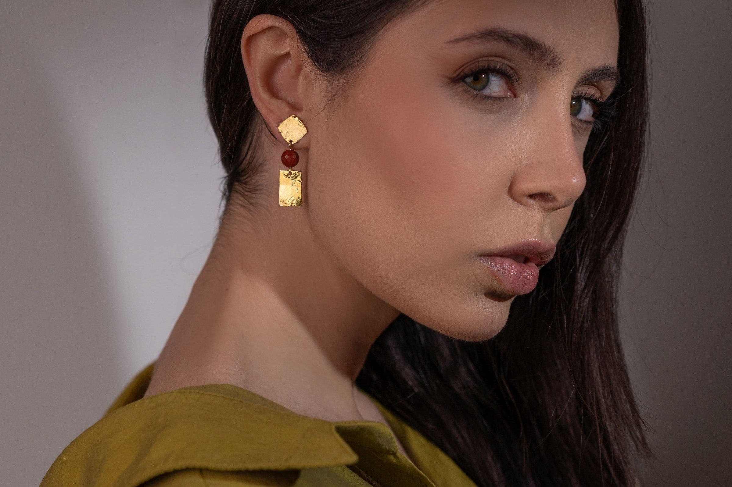 Petites boucles d'oreilles puces en métal martelé doré et perles de Mokaïte acajou, de forme losanges et rectangles, portées à l'oreille par une femme brune