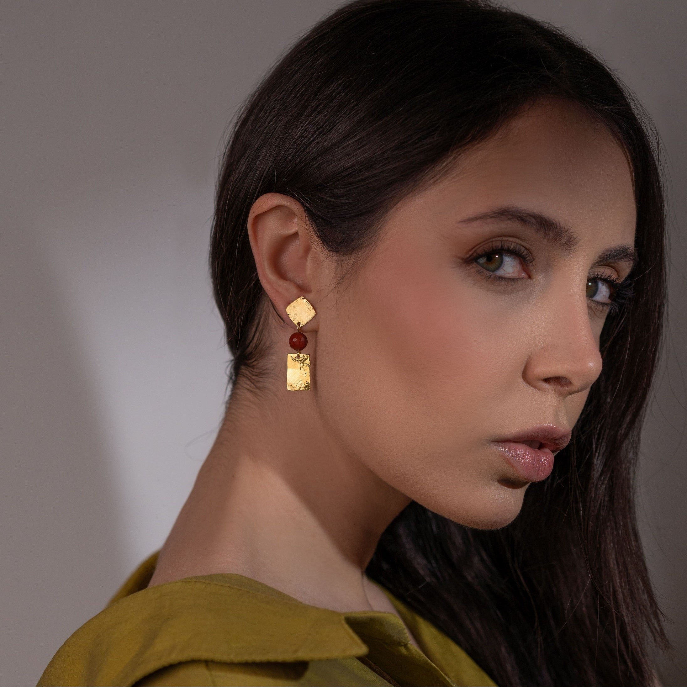 Petites boucles d'oreilles puces en métal martelé doré et perles de Mokaïte acajou, de forme losanges et rectangles, portées à l'oreille par une femme brune