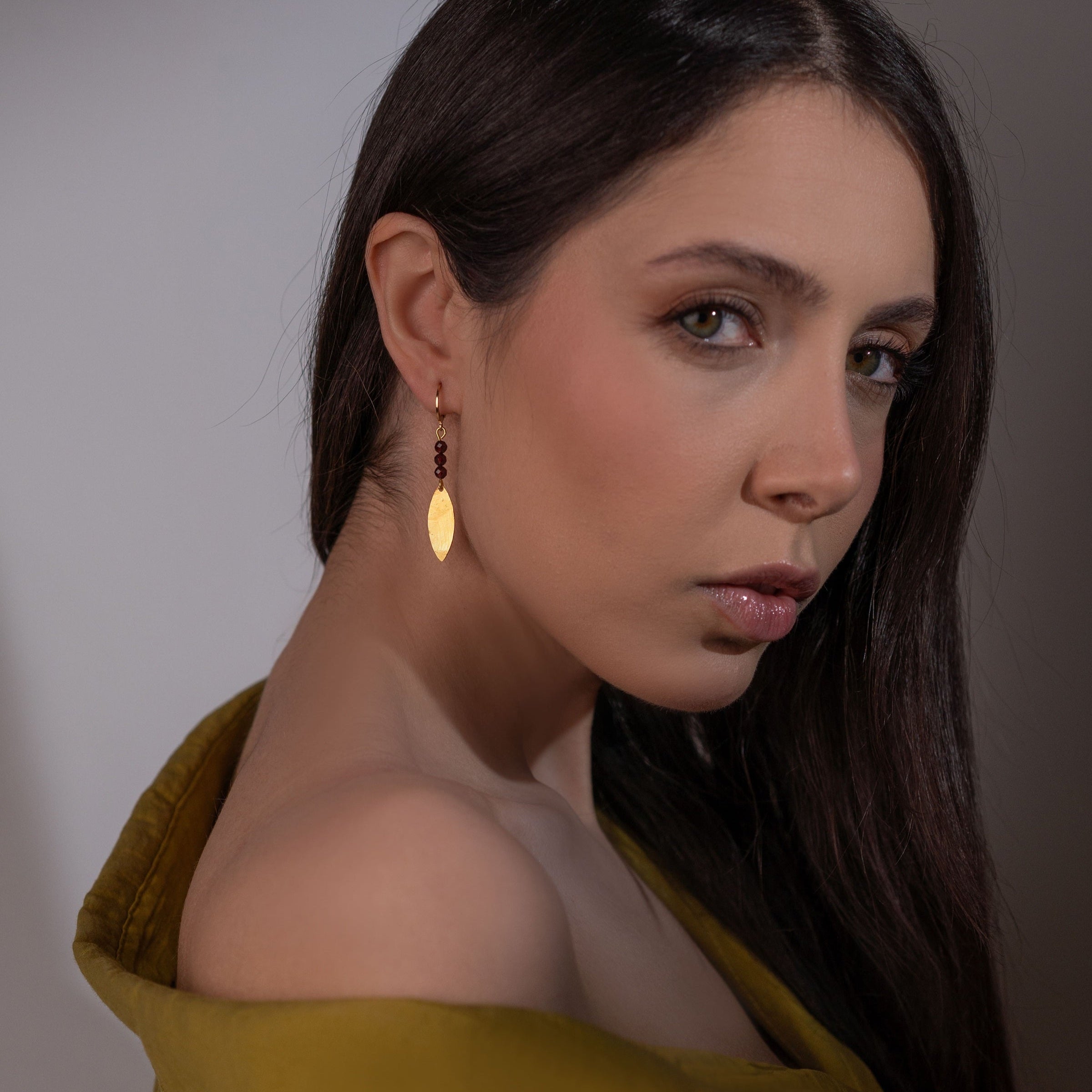 Boucles d'oreilles fines en pointe en métal martelé doré à l'or fin et petites perles de grenat, portées à l'oreille par une femme brune