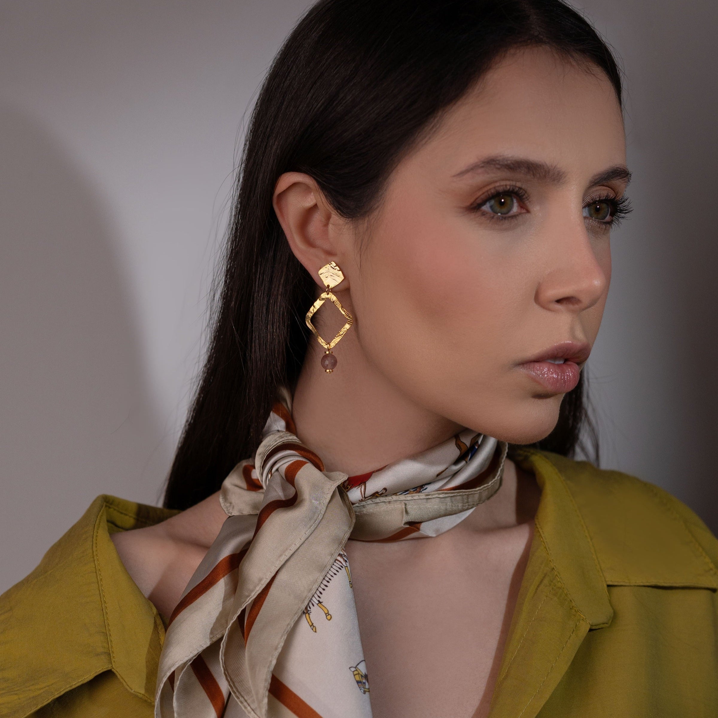 Boucles d'oreilles losanges montées sur puces en métal martelé doré à l'or fin et perles de quartz fraise, portées à l'oreille par une femme brune