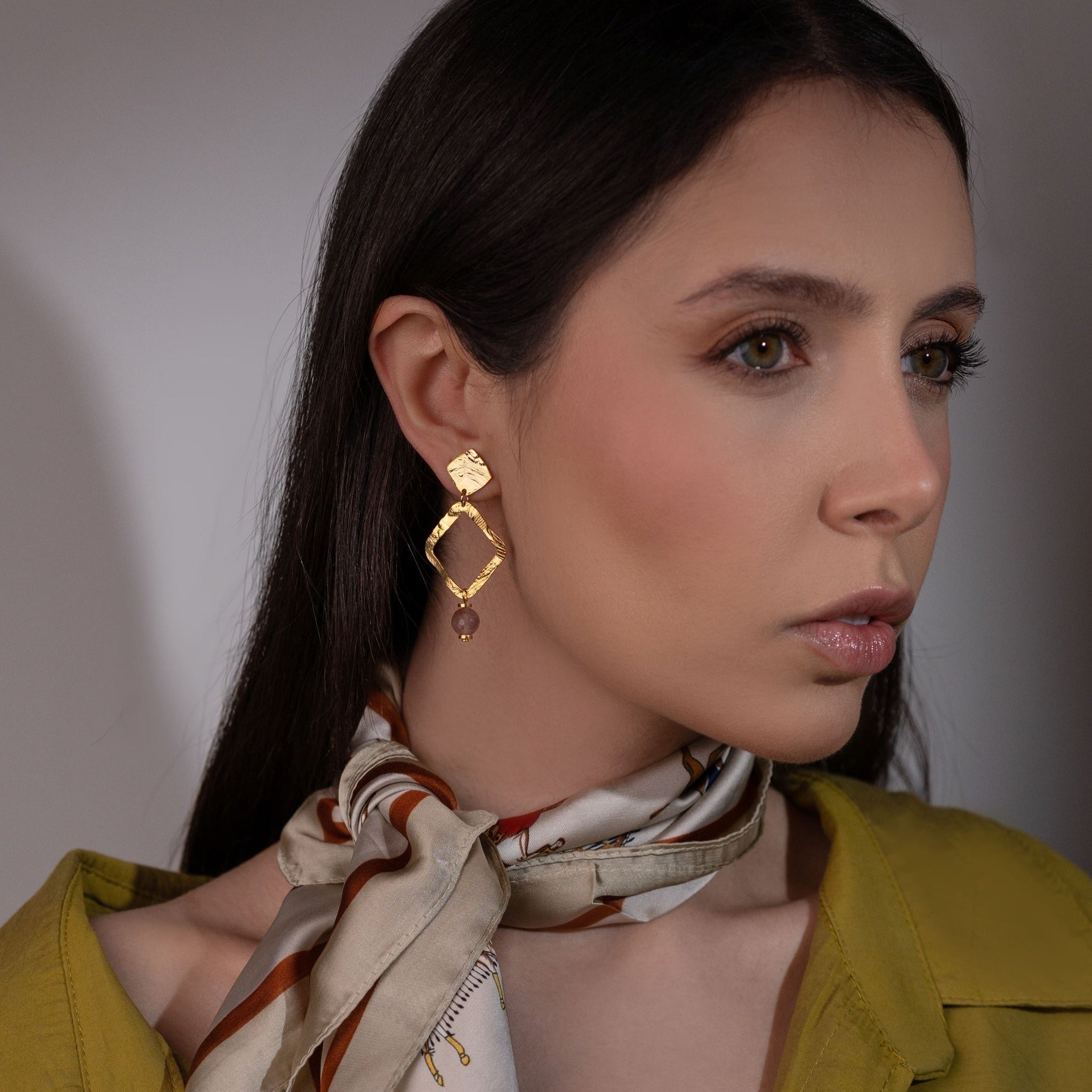 Boucles d'oreilles losanges montées sur puces en métal martelé doré à l'or fin et perles de quartz fraise, portées à l'oreille par une femme brune