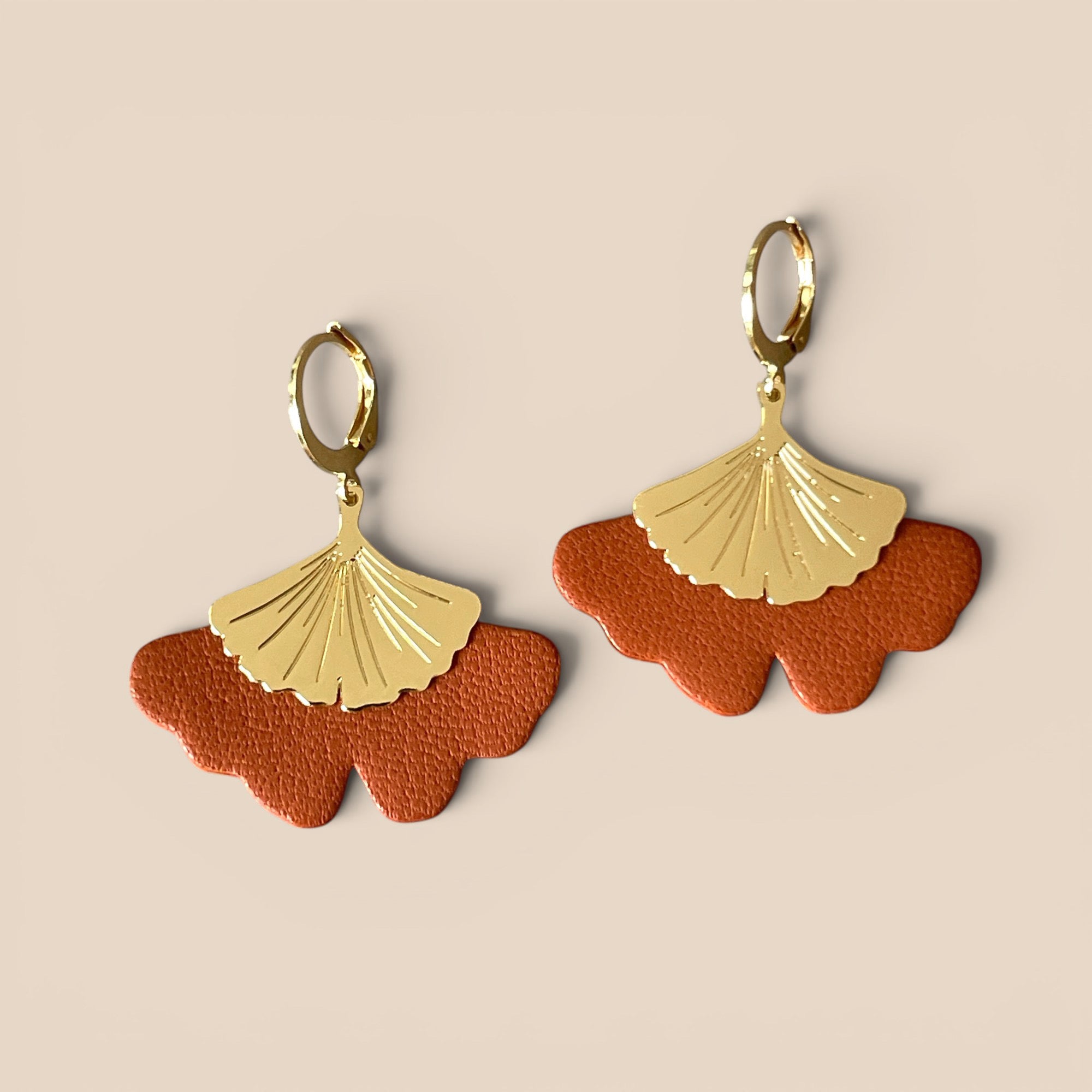 Boucles d'oreilles dorées pendantes en cuir terracotta en forme de gingko, sur fond beige