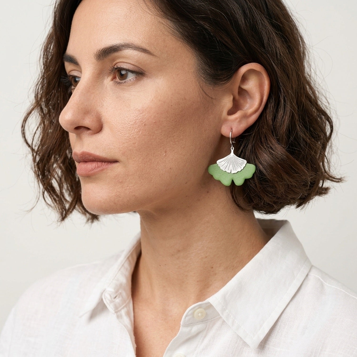 Boucles d'oreilles argentées pendantes en cuir vert pomme pastel en forme de gingko, portées aux oreilles par une femme brune