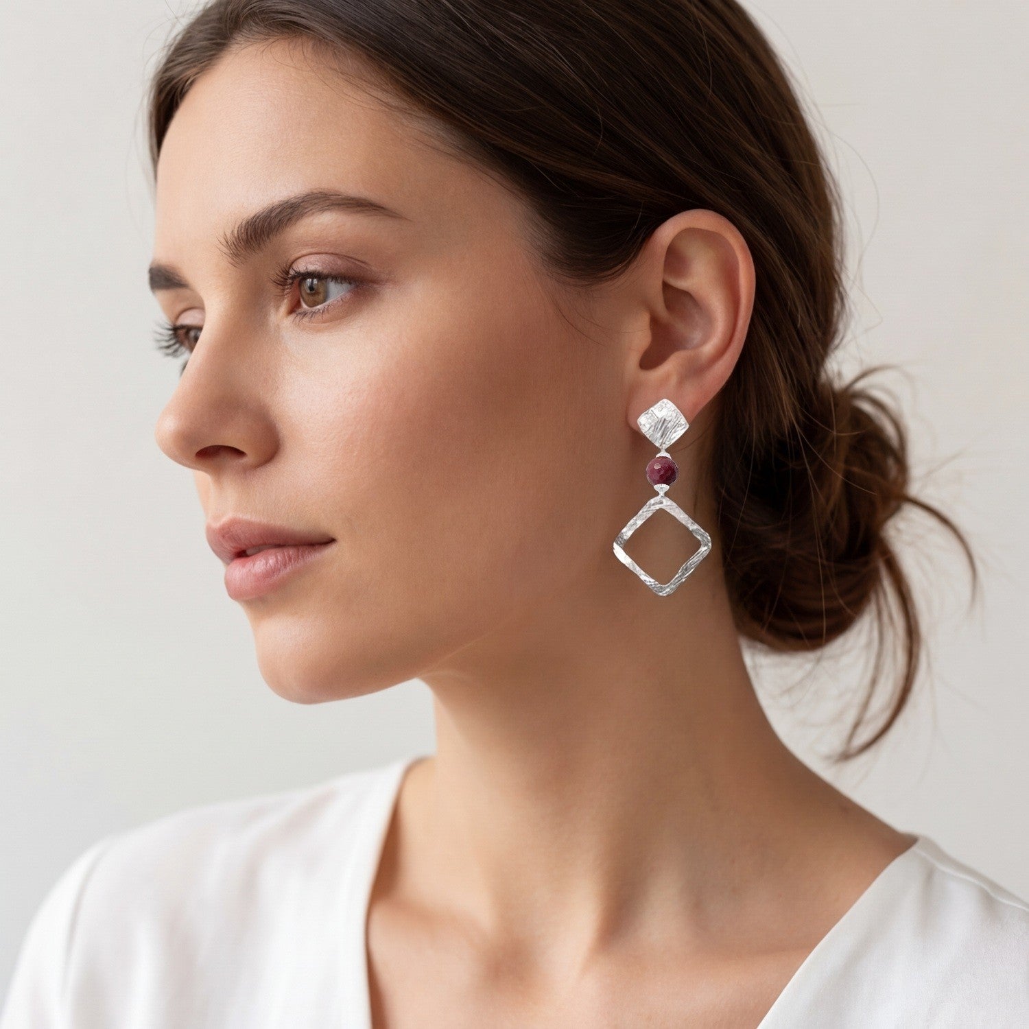 Boucles d'oreilles losanges sur puces en métal martelé argenté et perles de Mokaïte bordeaux, portées à l'oreille par une femme brune