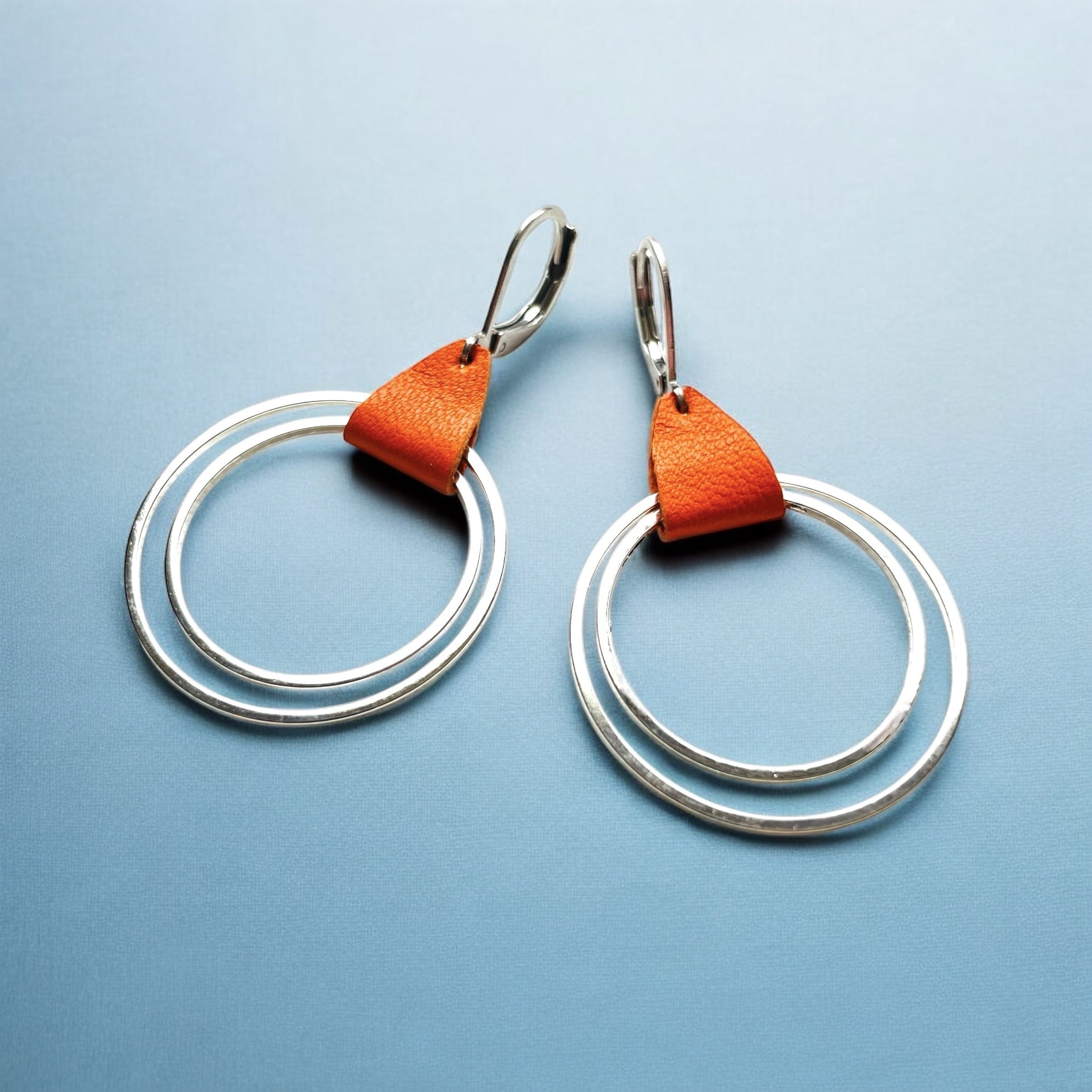 Créoles argentées du modèle Vega de la marque Nune, avec une touche de cuir orange, sur fond bleuté