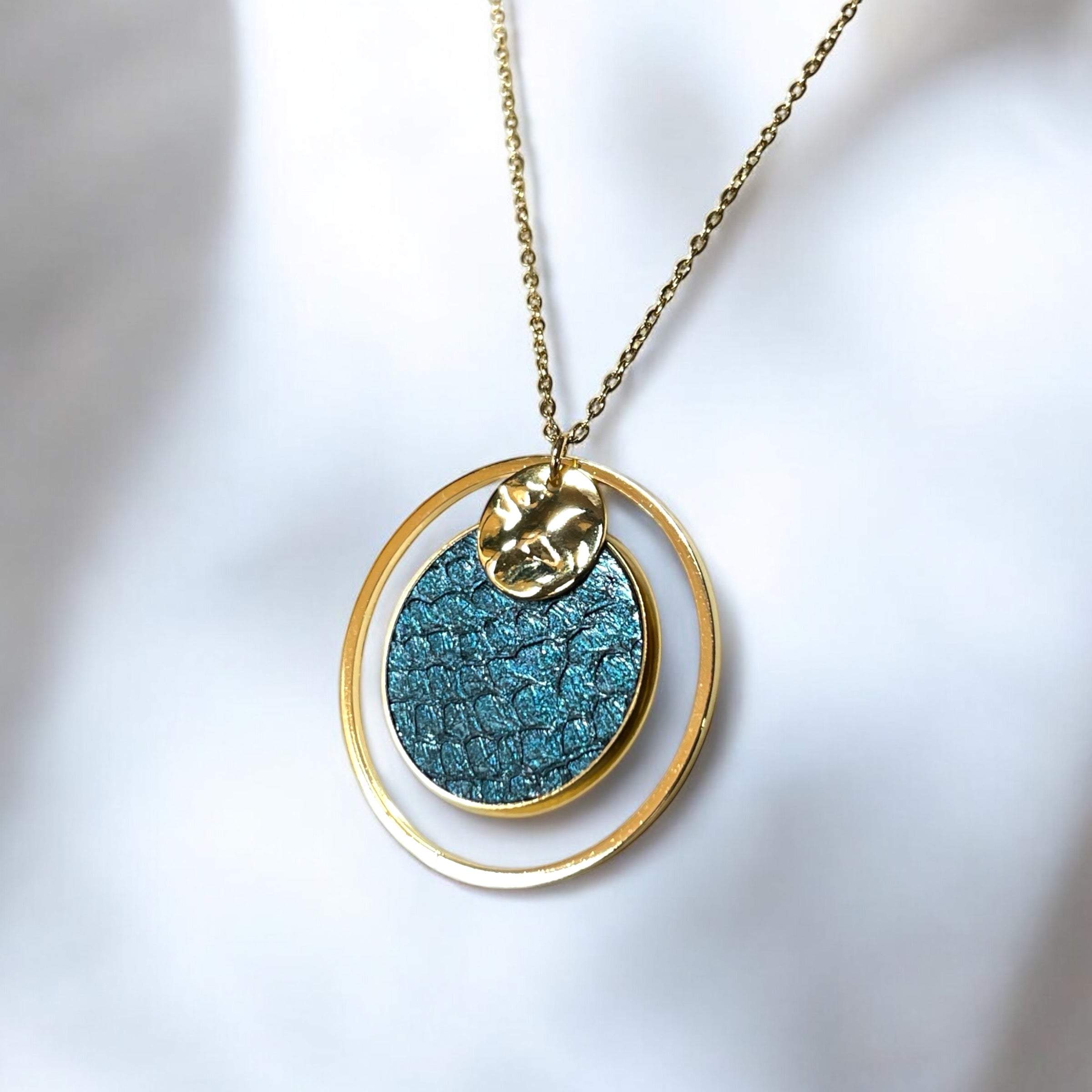 Fin collier doré avec pendentif rond en anneau avec une pièce de cuir marin bleu paon métallisé, sur fond blanc