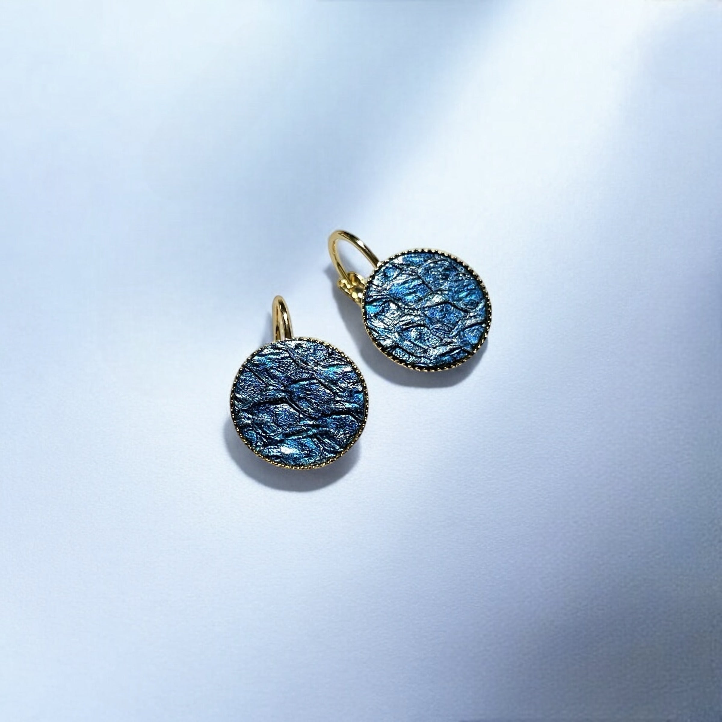 Petites boucles d'oreilles dormeuses dorées serties de fin cuir marin saphir métallisé, sur fond bleu