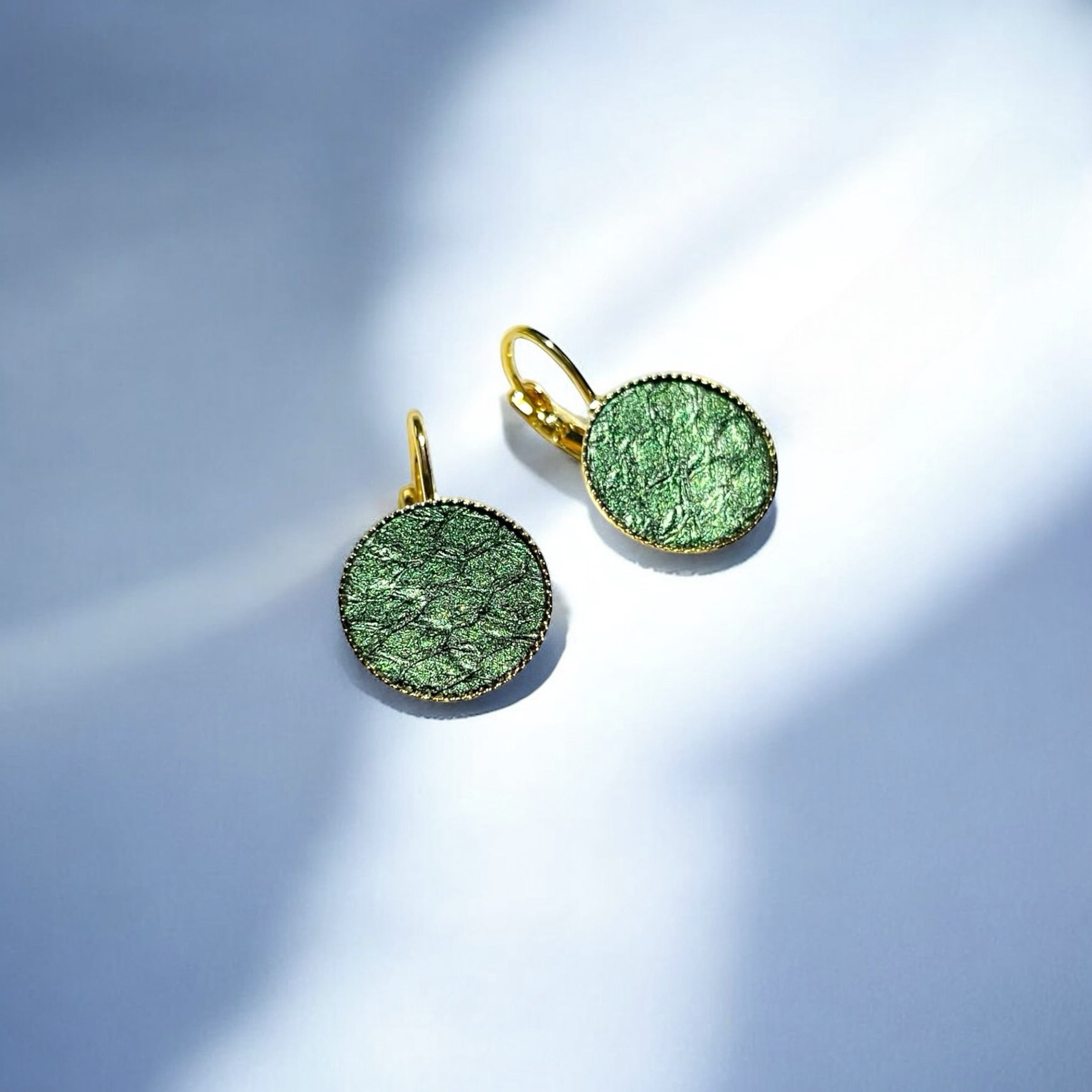 Petites boucles d'oreilles dormeuses dorées serties de fin cuir marin vert bouteille métallisé, sur fond bleu