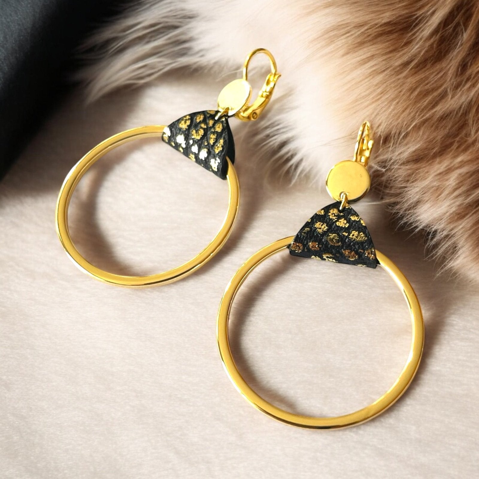 Boucles d'oreilles dorées, grands anneaux créoles, noués par un morceau de cuir noir grainé or aux accroches dormeuses, présentées sur fond beige avec fourrure