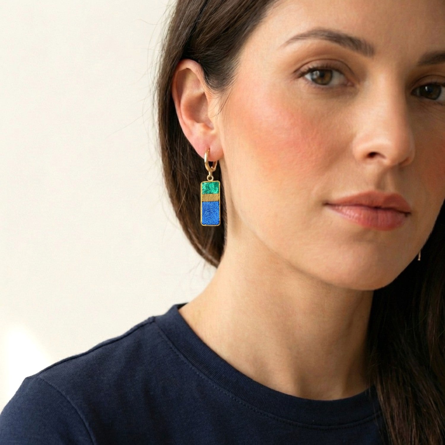 Petites boucles d'oreilles dorées pendantes rectangulaires verticales tricolores en cuirs émeraude métallisé, bronze doré et bleu cobalt métallisé, portées au oreilles par une femme brune