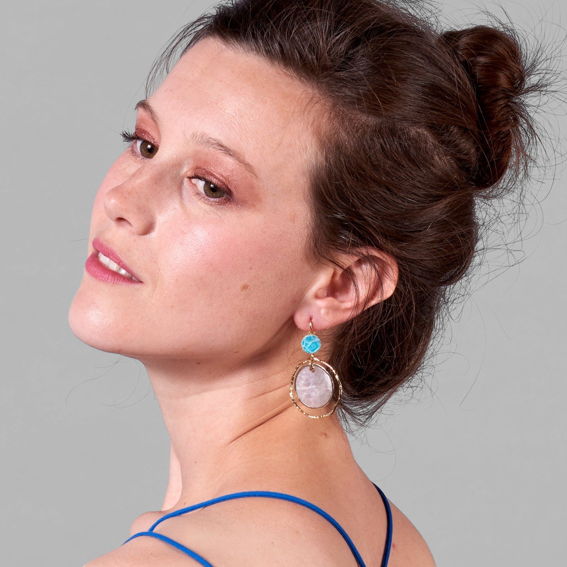Jeune femme à chignon vue de profil de près portant les boucles d'oreilles dorées Nune modèle Bali Créoles Capiz en cuir turquoise sur fond gris