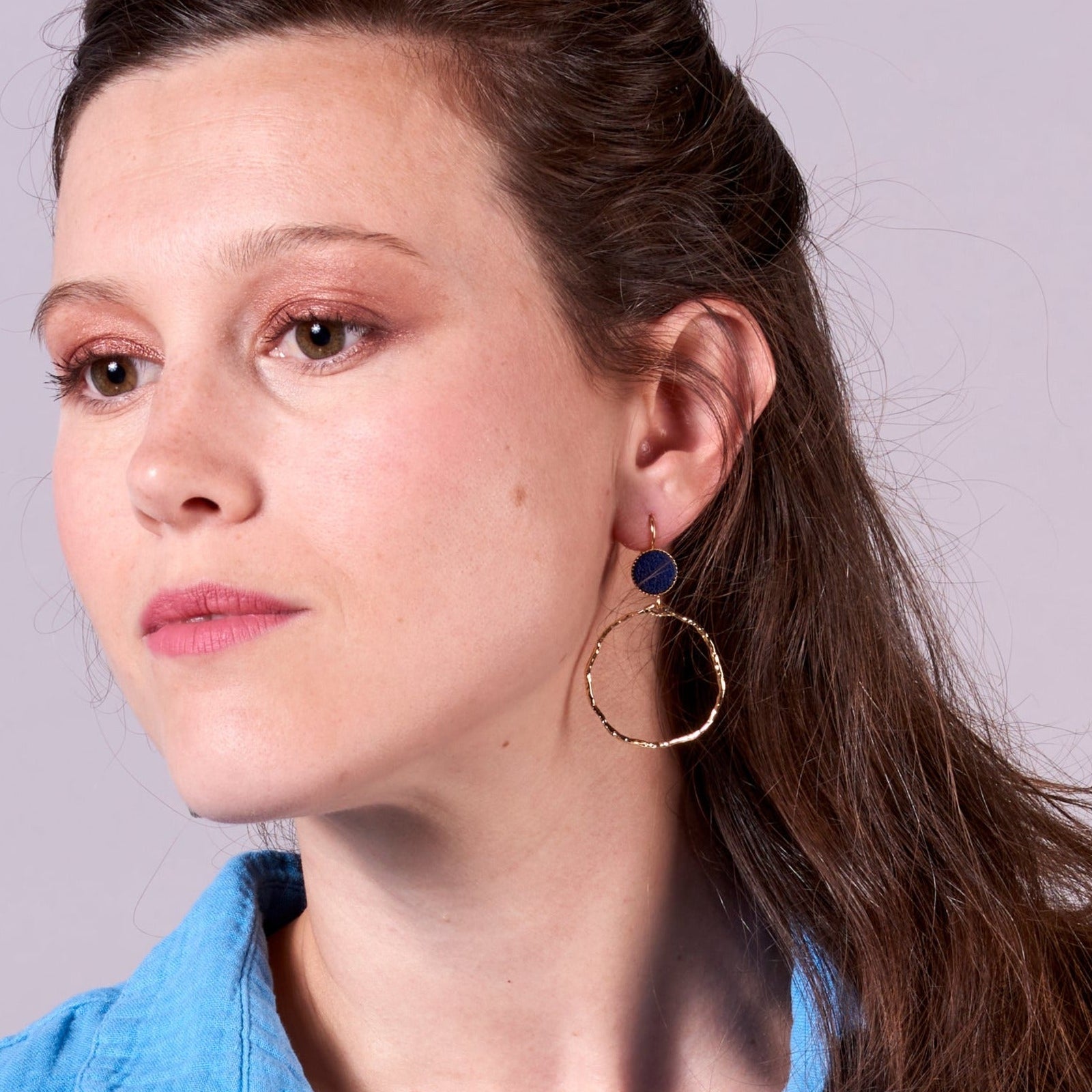 Jeune femme vue de 3/4 portant les boucles d'oreilles dorées Nune modèle Bali Créoles en cuir marine satiné, sur fond gris