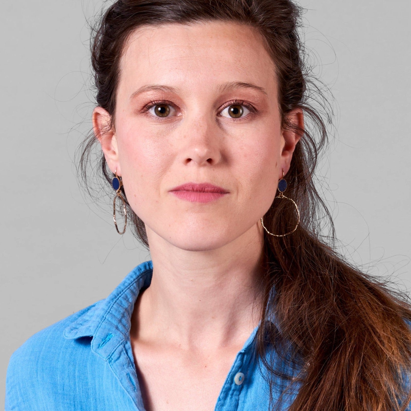 Jeune femme vue de face portant les boucles d'oreilles dorées Nune modèle Bali Créoles en cuir marine satiné, sur fond gris