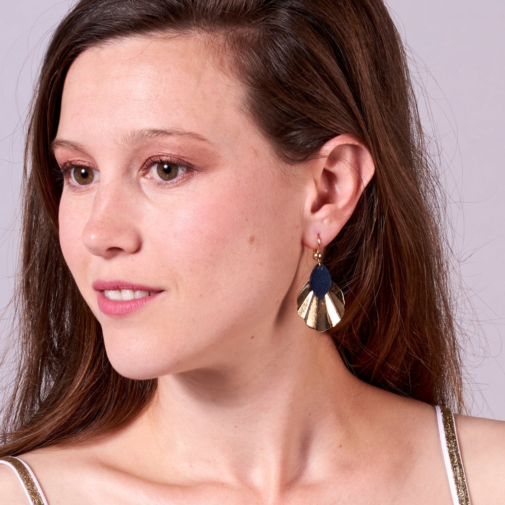 Jeune femme vue de prÚs portant les Boucles d'oreilles dorées Nune modÚle Okita en forme de petits coquillages avec pétales de cuir bleu marine satiné, sur fond mauve