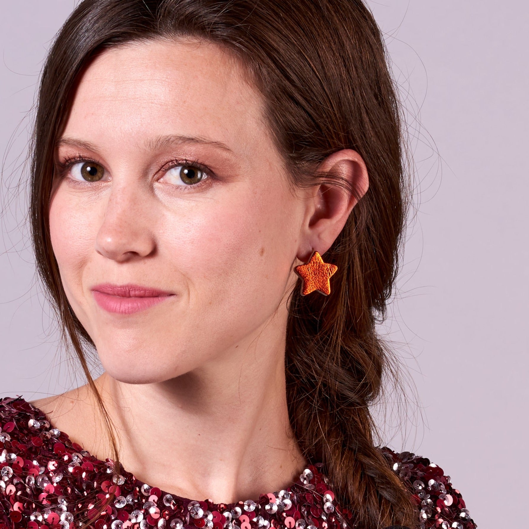 Jeune fille aux longs cheveux bruns vue de 3/4 portant les boucles d'oreilles Diva de Nune en forme d'étoile en cuir recyclé orange sanguine métallisé, sur mauve