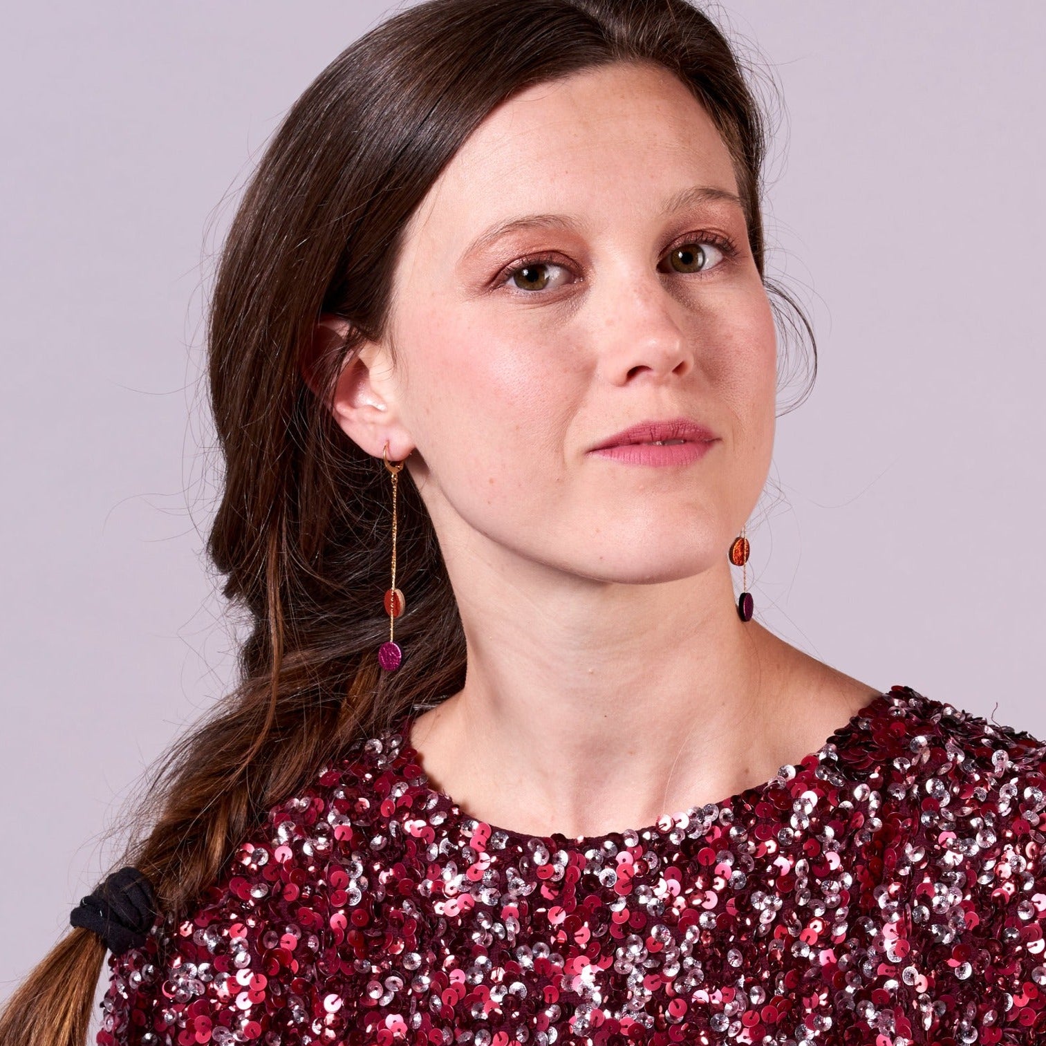 Jeune fille vue de loin portant les Longues et fines boucles d'oreilles Nune dorées terminées petits disques de cuir recyclé orange sanguine métallisé et fushia métallisé. Modèle Merry