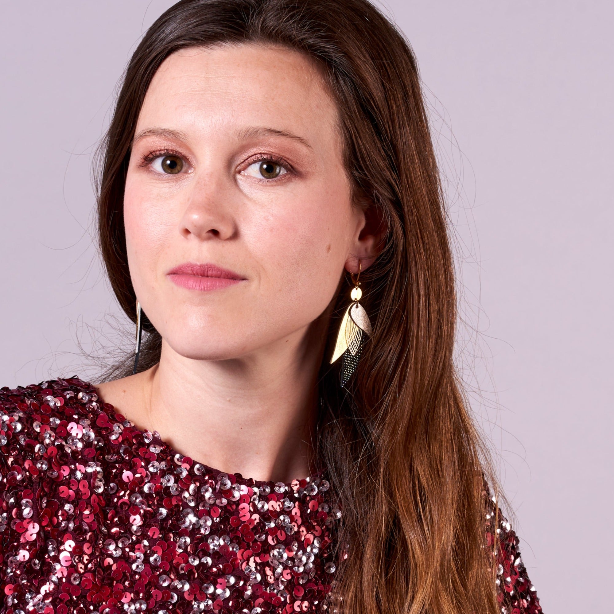 Jeune femme aux longs cheveux bruns portant les boucle d'oreille dorée Nune modèle Ara en plumes de cuir argent, doré brillant et noir grainé irisé, sur fond mauve