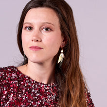 Charger l'image dans la galerie, Jeune femme aux longs cheveux bruns portant les boucle d'oreille dorée Nune modèle Ara en plumes de cuir argent, doré brillant et noir grainé irisé, sur fond mauve