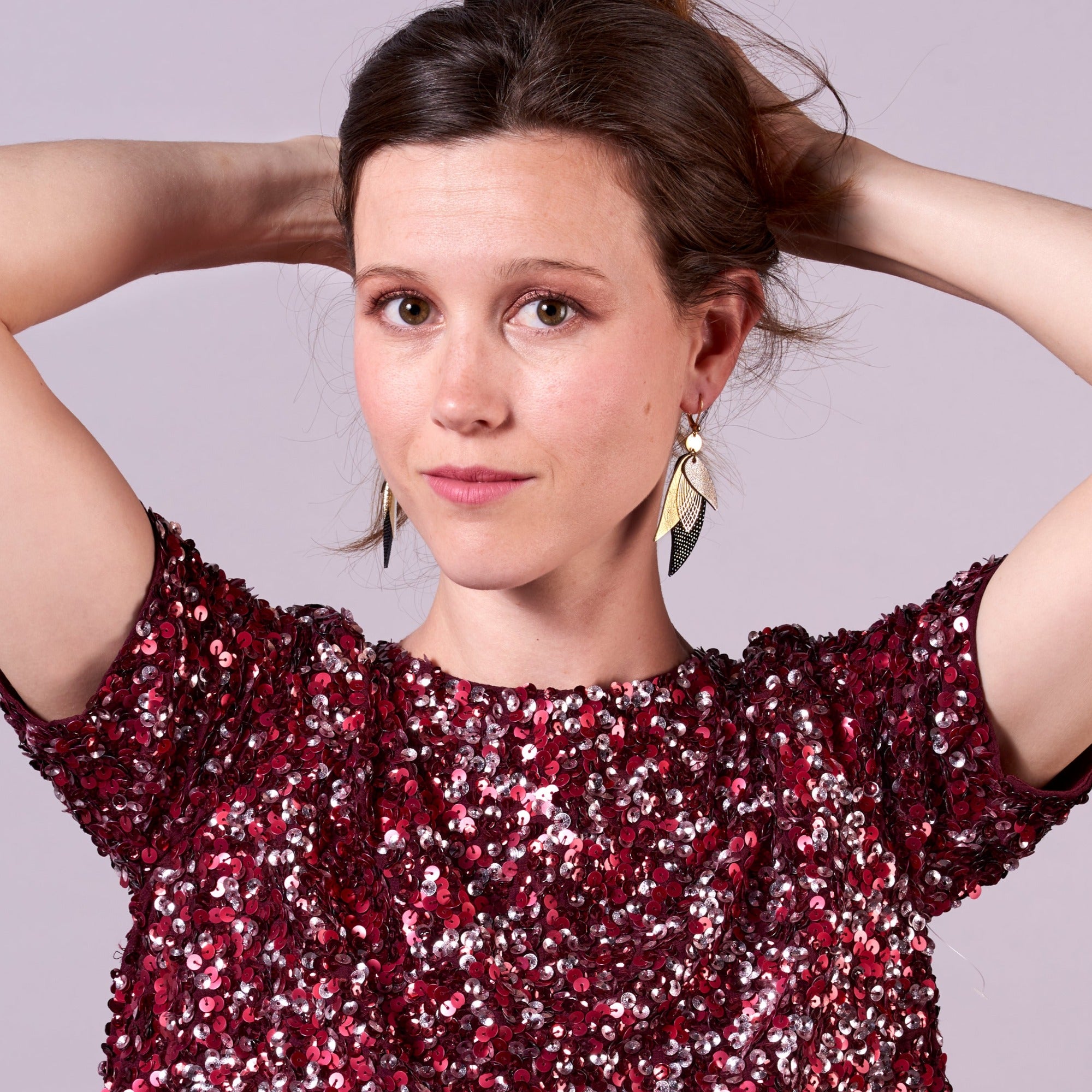 Jeune femme tenant ses cheveux bruns portant les boucle d'oreille dorée Nune modèle Ara en plumes de cuir argent, doré brillant et noir grainé irisé, sur fond mauve