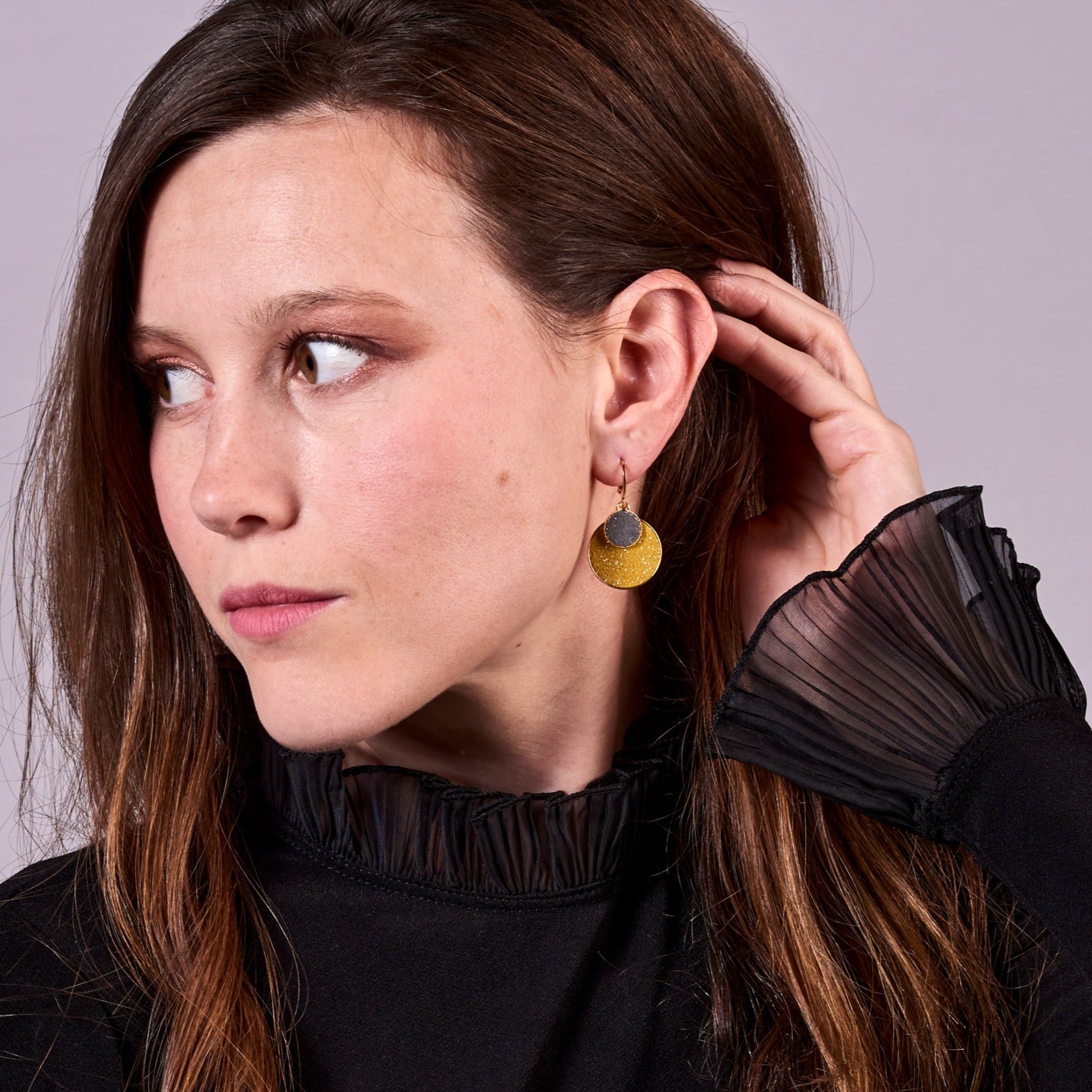 Jeune femme brune vue de près de profil portant les Petites boucles d'oreilles dorées rondes Bulles Pailletées de Nune avec doubles sertis de cuirs pailletés safran et gris, sur fond mauve