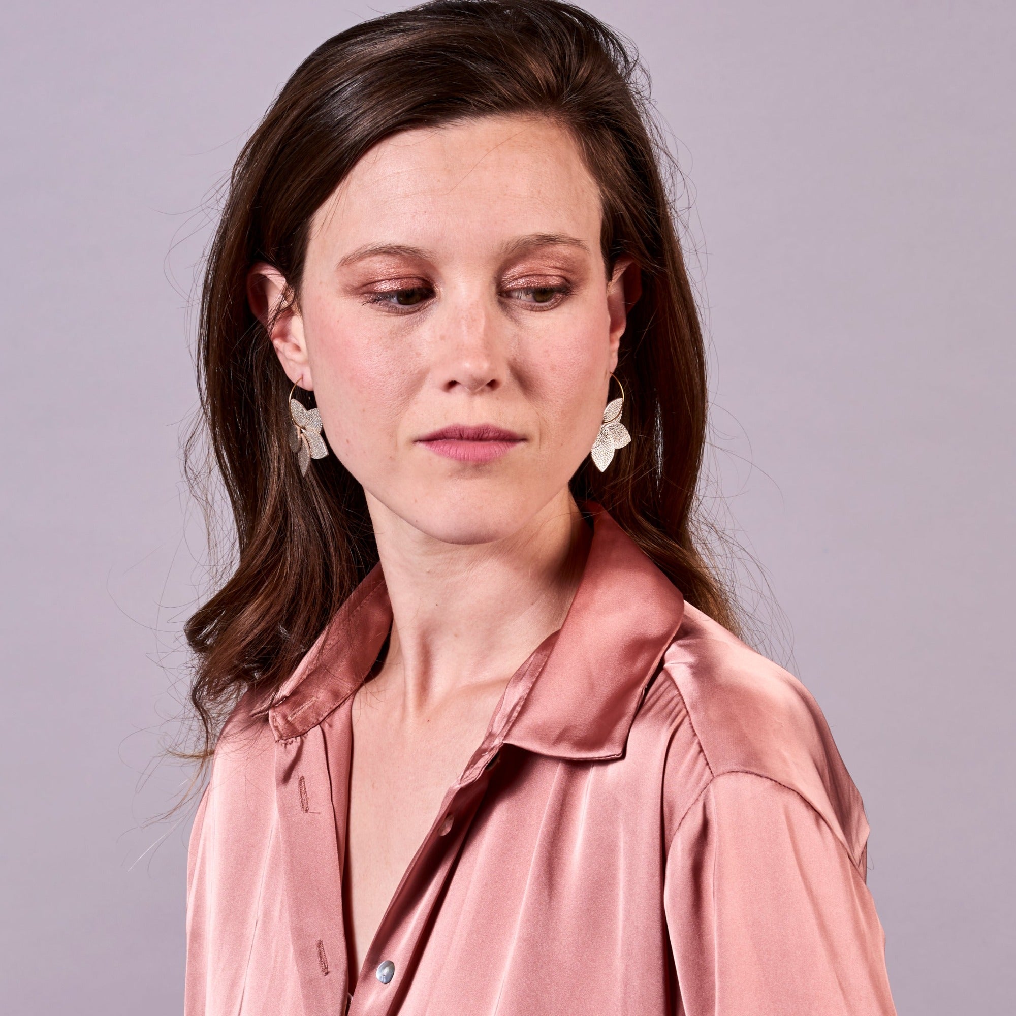Jeune femme vue de face et en buste portant les petites créoles Jasmin en pétales de cuir fushia métallisé, en forme de fleurs, sur fond mauve