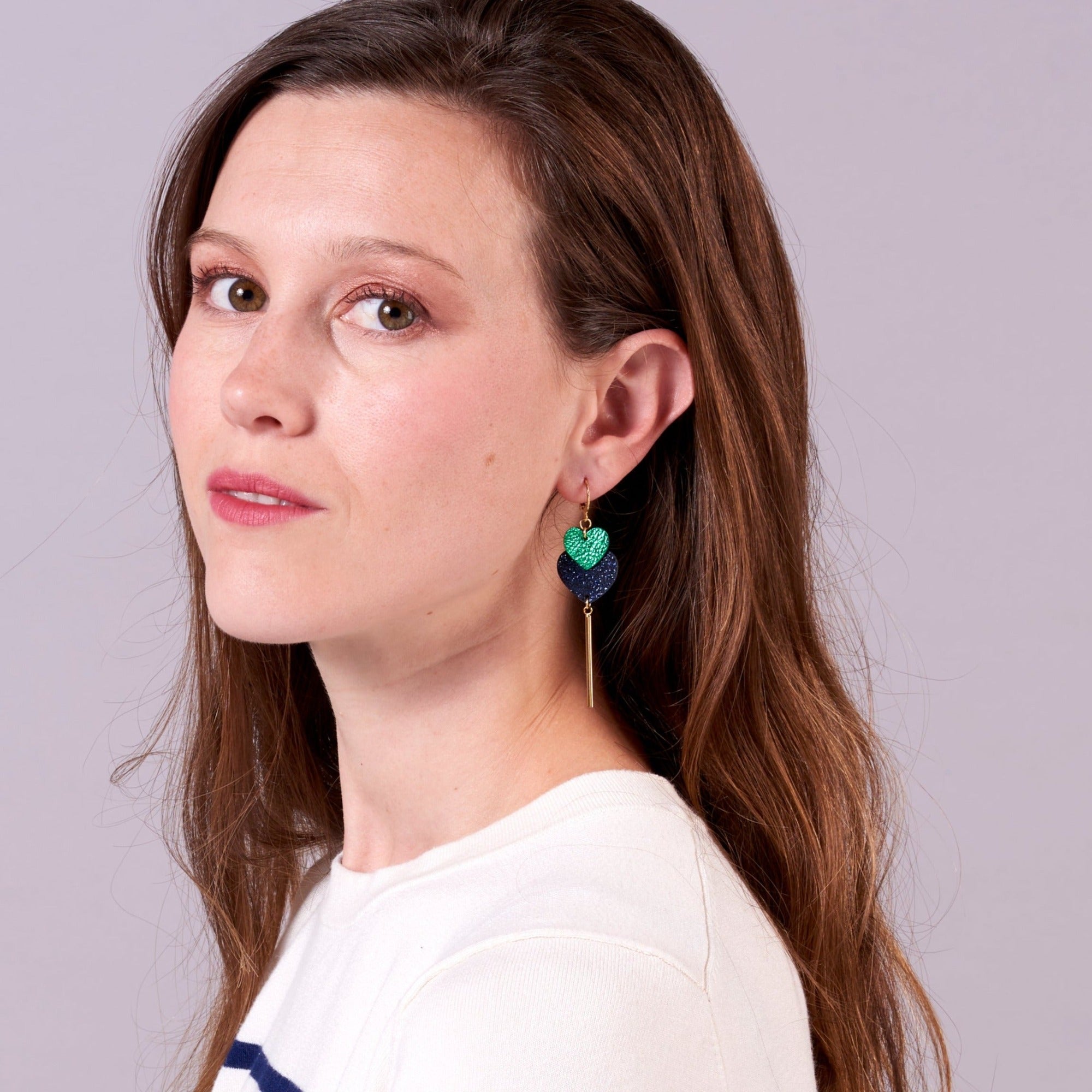 Jeune femme vue de profil portant les Longues boucles d'oreilles dorées modèle Félicie avec 2 cœurs imbriqués en cuirs recyclés vert métallisé et bleu marine pailleté, sur fond mauve