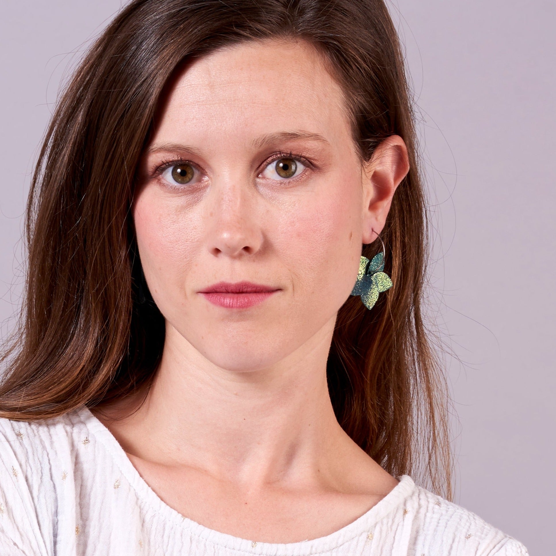 Jeune femme vue de face et de près portant les petites créoles Jasmin Argenté en pétales de cuir vert canard pailleté irisé, en forme de fleurs, sur fond mauve