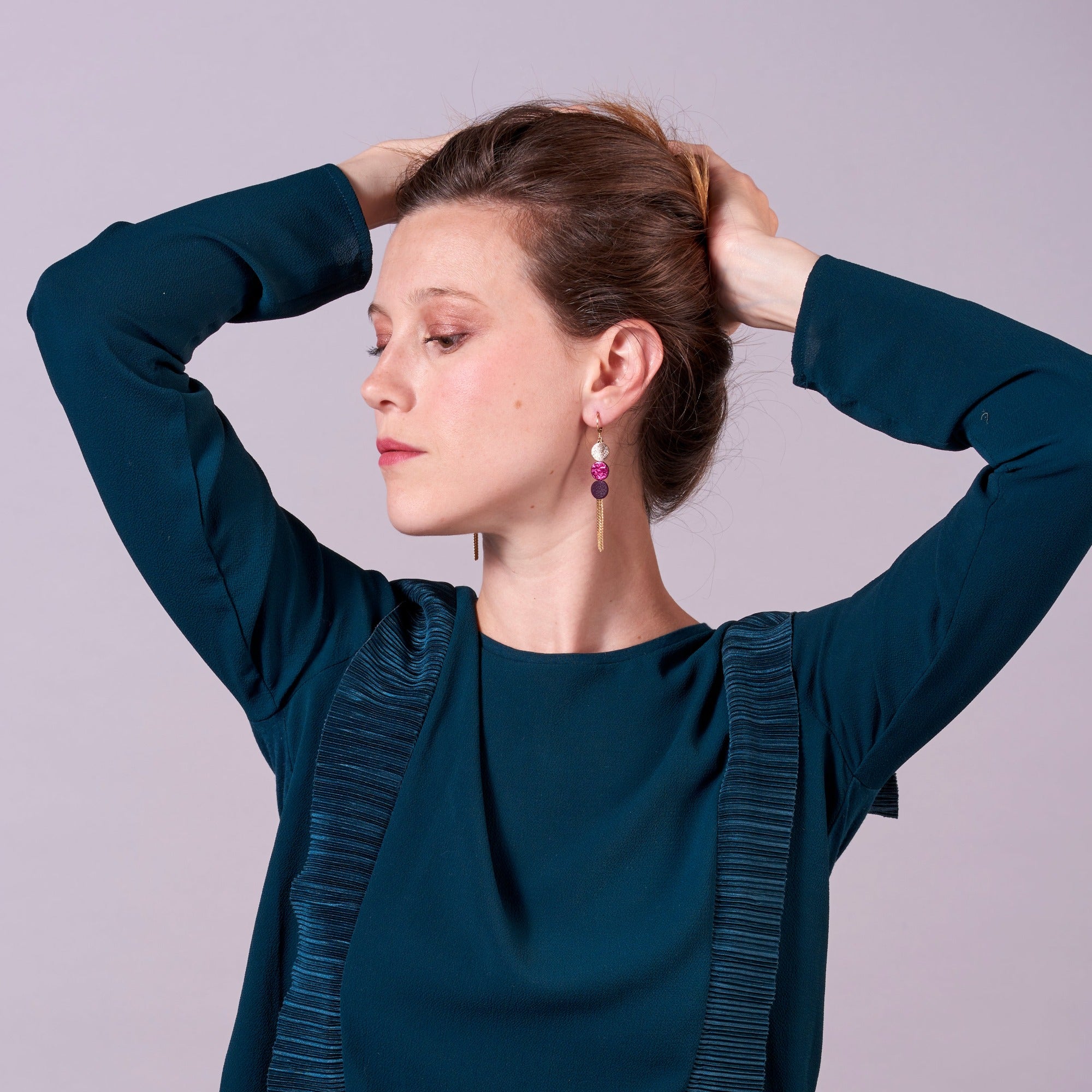 Jeune fille vue en buste portant les longues et fines boucles d'oreilles Janis de Nune, boucles d'oreilles à franges dorées avec 3 petits disques de cuir recyclé cannelle dorée satinée, fuchsia métallisé et violine satinée sur fond mauve