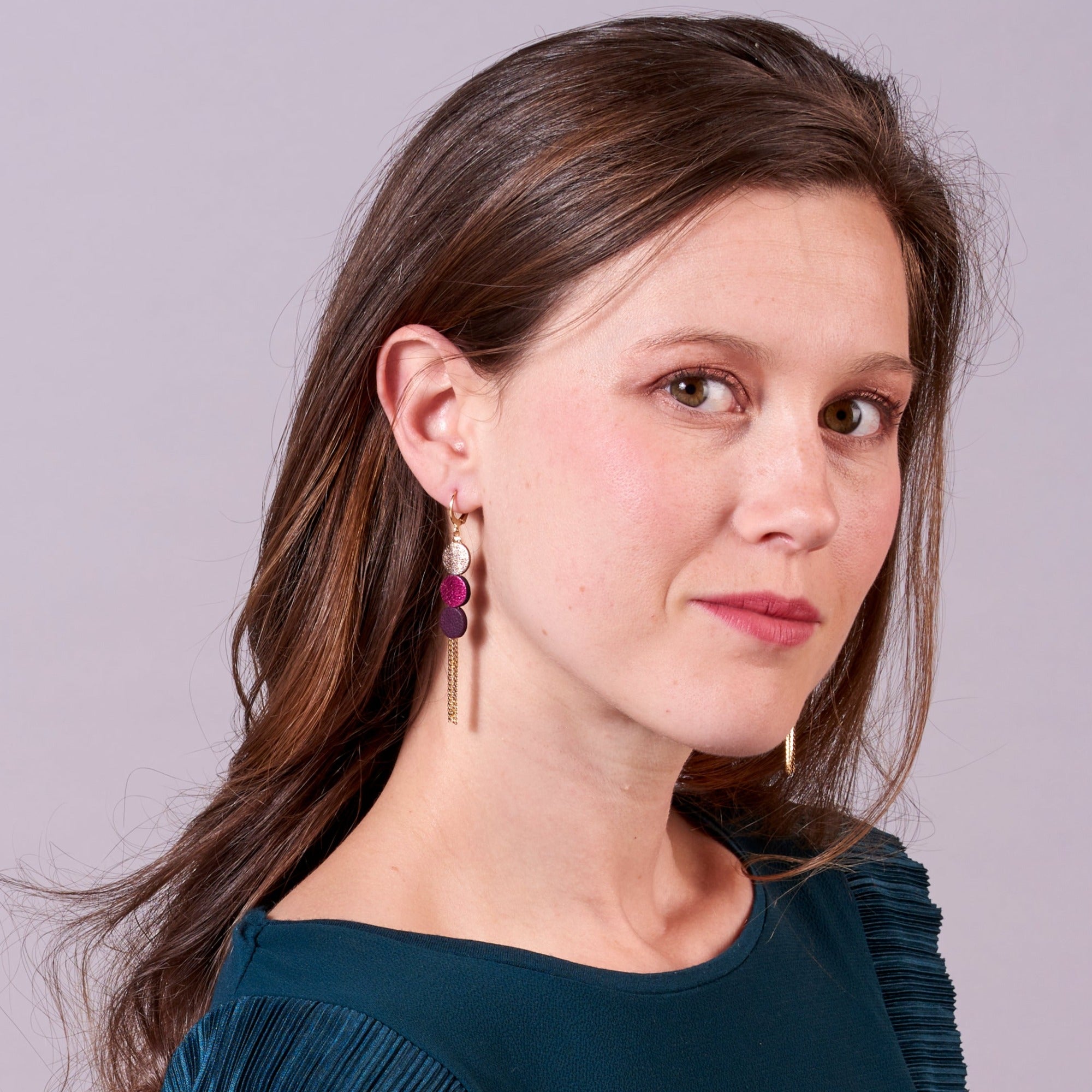 Jeune fille vue de près portant les longues et fines boucles d'oreilles Janis de Nune, boucles d'oreilles à franges dorées avec 3 petits disques de cuir recyclé cannelle dorée satinée, fuchsia métallisé et violine satinée sur fond mauve