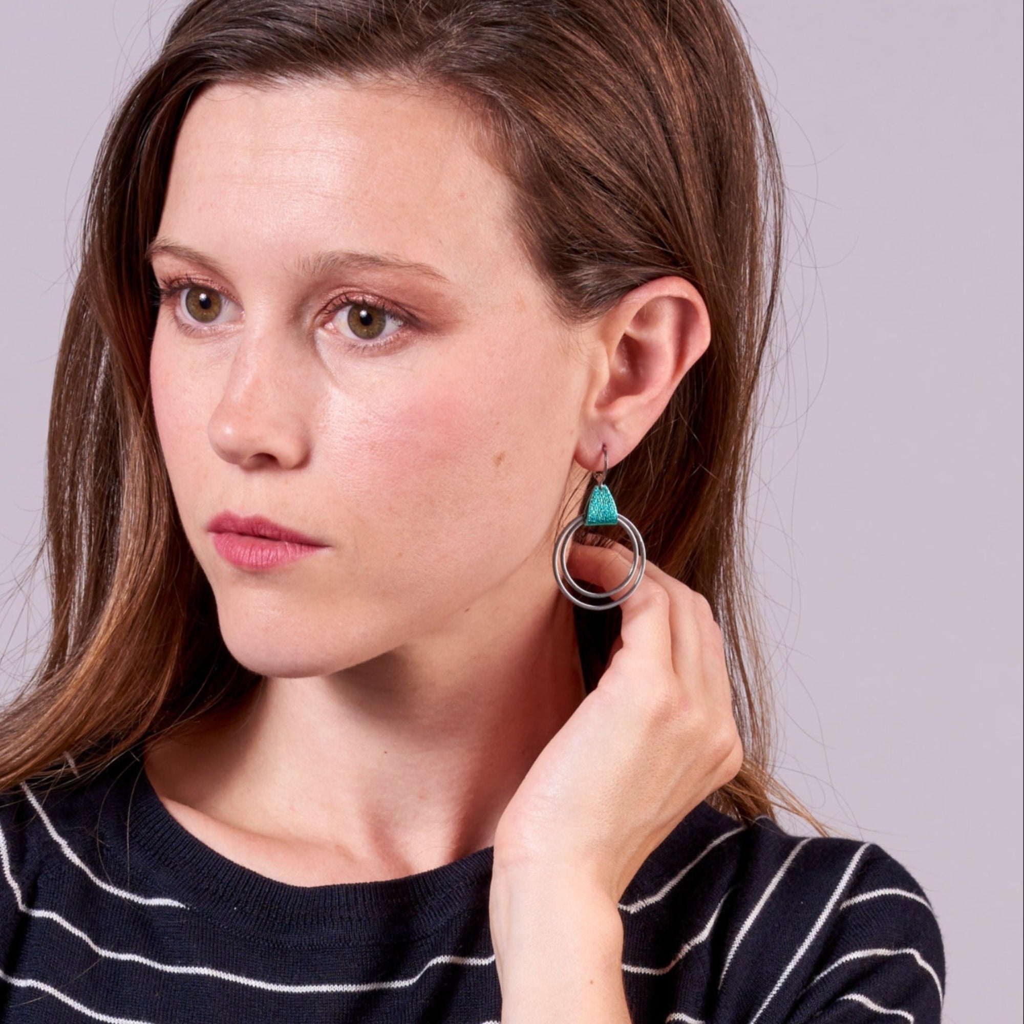 Jeune femme portant les Créoles argentée du modèle Vega de la marque Nune, avec une touche de cuir turquoise métallisé, sur fond mauve