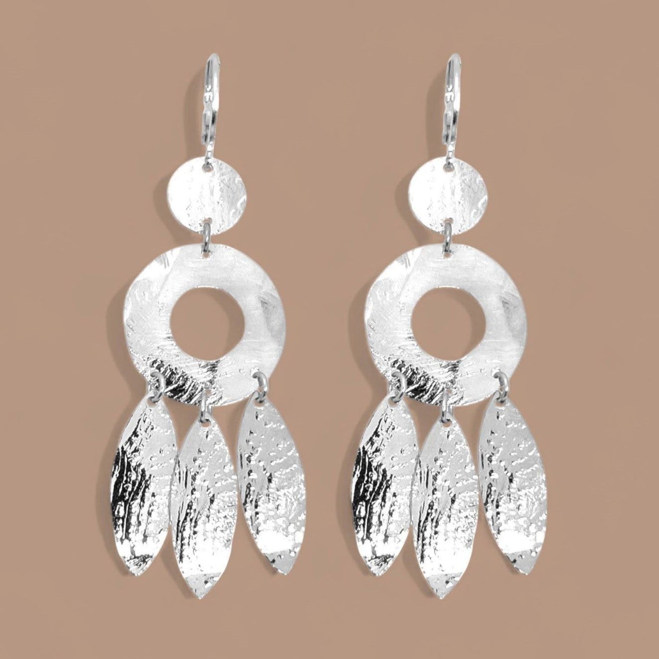 Longues boucles d'oreilles argentées en métal martelé, de style ethnique bohème chic, avec leurs pampilles en forme de plumes, sur fond beige
