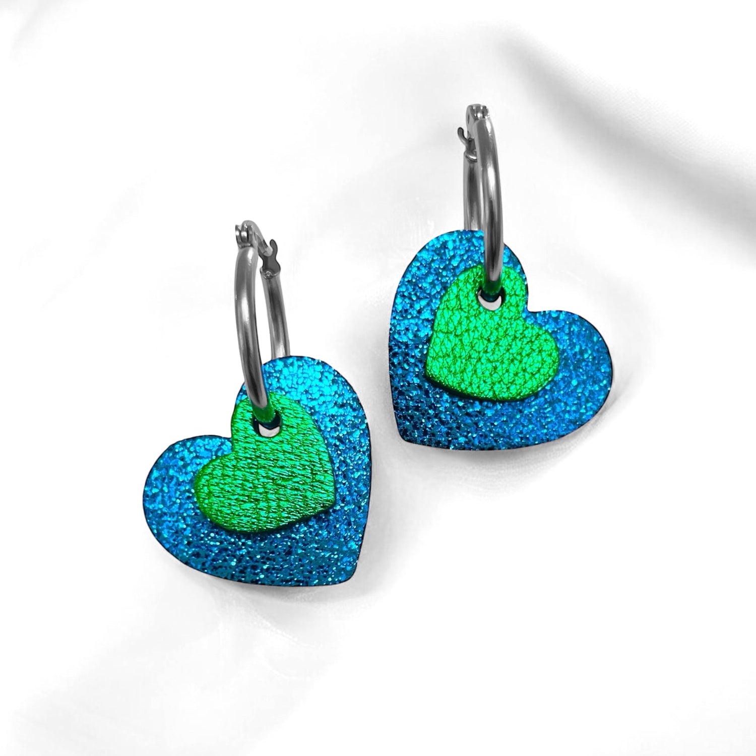 Boucles créoles argentées avec pendentifs en forme de cœurs bleu roi métallisé et vert métallisé, posées sur fond blanc