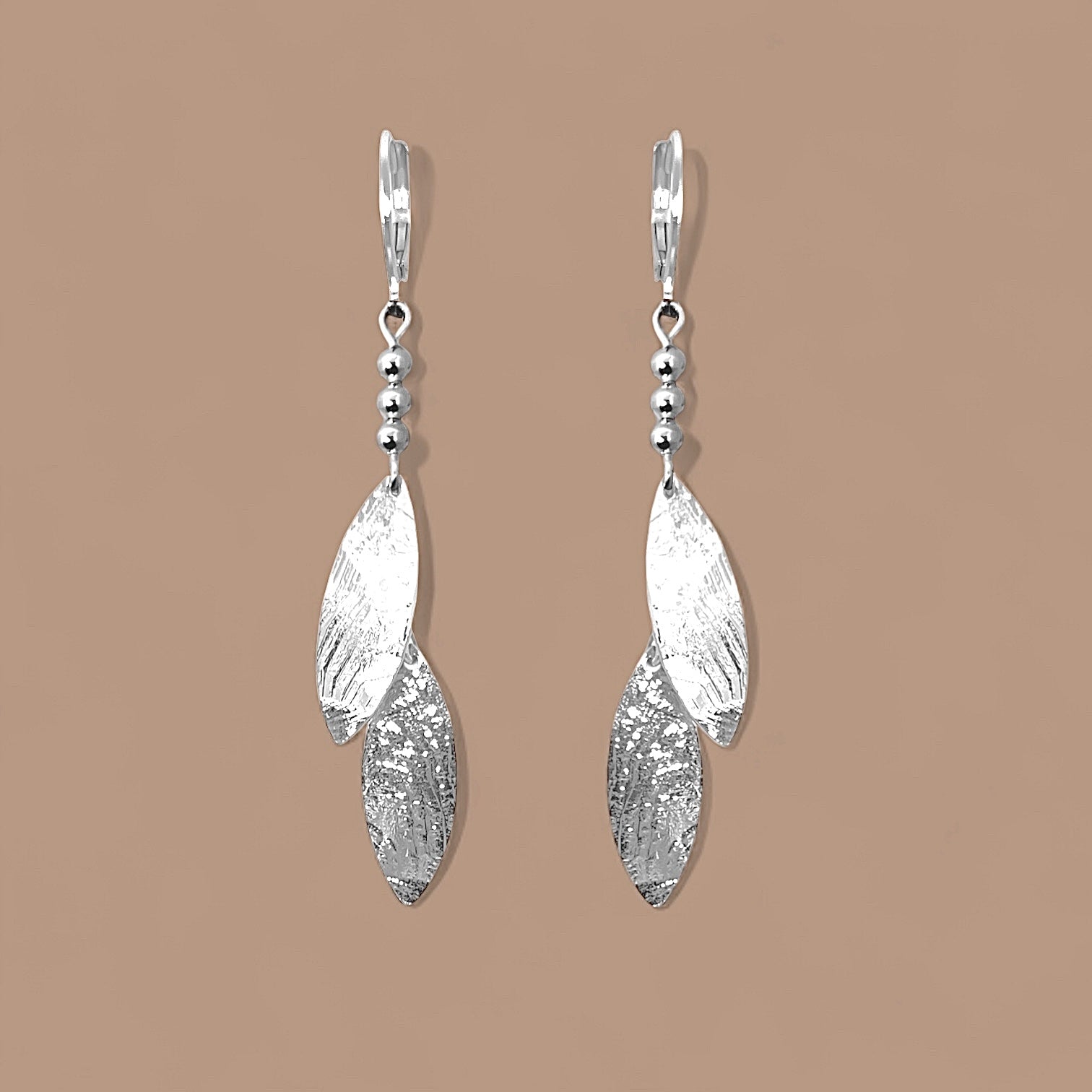Longues boucles d'oreilles fines avec double pointes en métal martelé argenté, sur fond beige