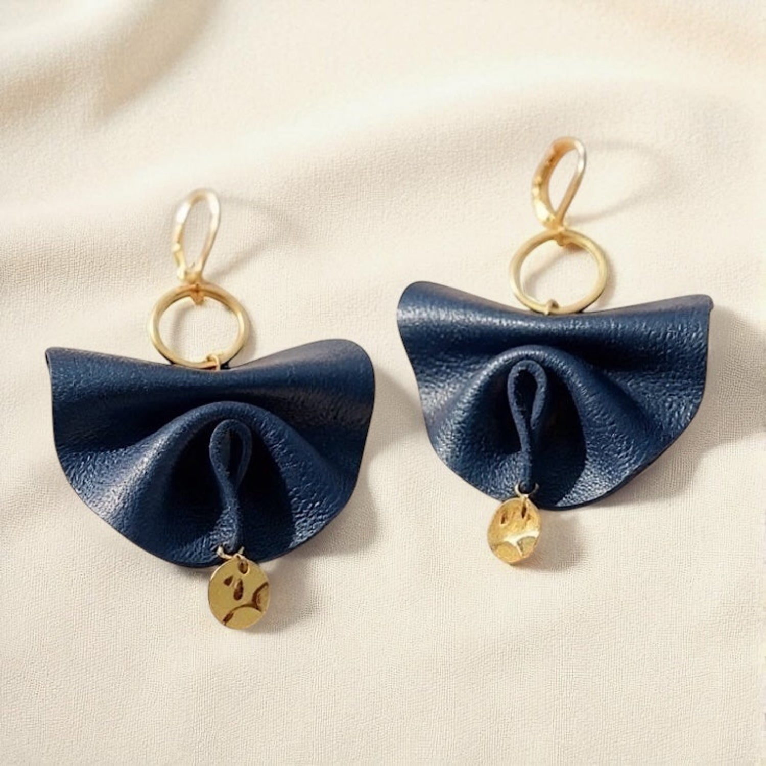 Grandes boucles d'oreilles dorées en éventails de cuir plissé recyclé bleu marine satiné, sur fond beige