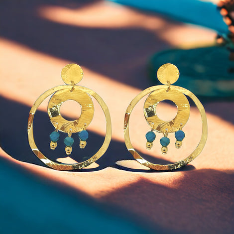 Grandes boucles d'oreilles puces créoles dorées pendantes longues rondes avec double anneau et petites perles en apatite bleue, modèle Bergame, sur fond bleu et orangé
